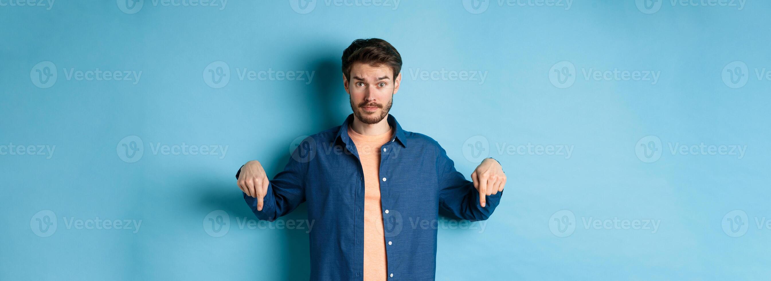Confused male model in casual clothes, looking puzzled and pointing fingers down, showing banner, standing on blue background photo