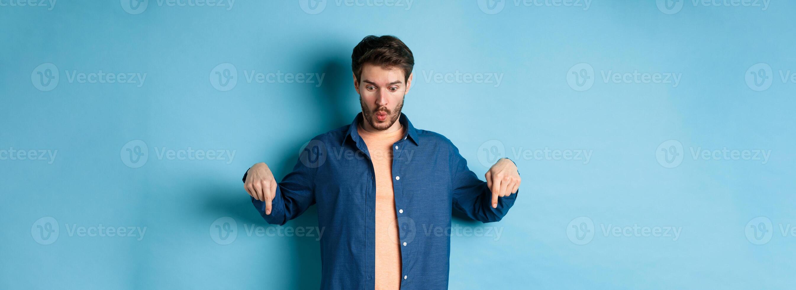 Impressed caucasian male model in casual clothes, say wow, looking and pointing fingers down at logo, standing on bllue background photo
