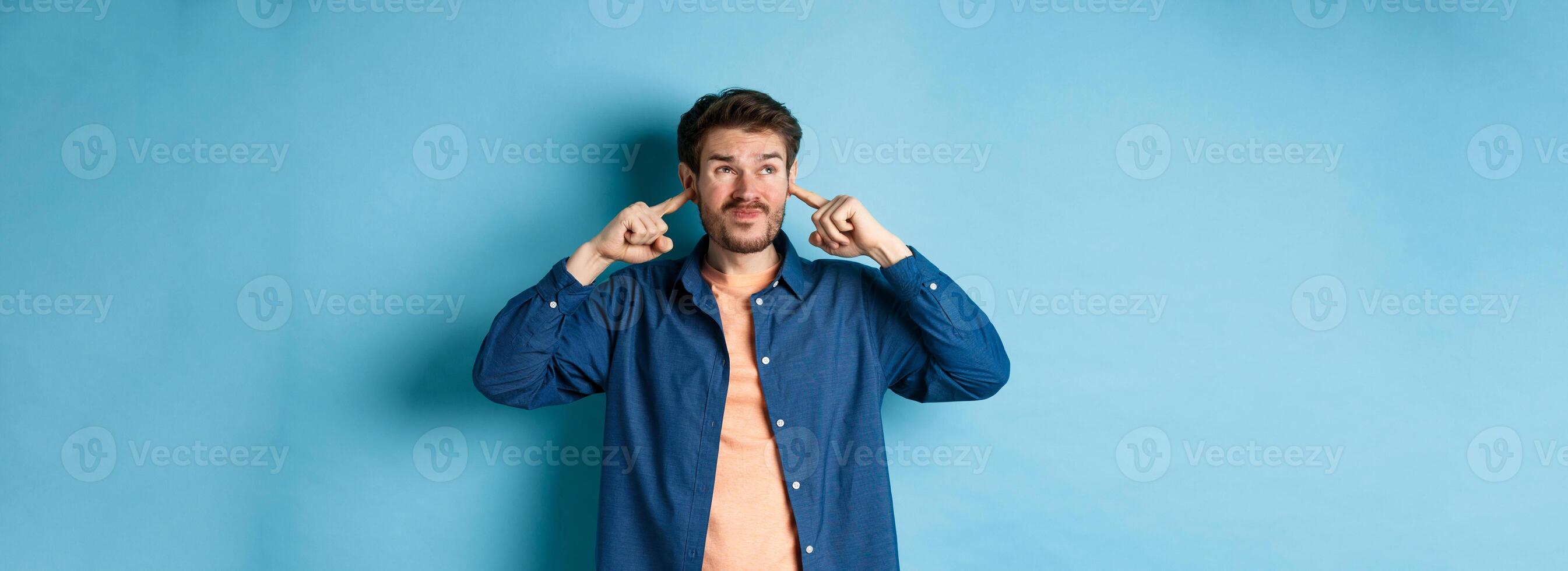Annoyed young man block sound, shut ears with fingers and grimacing, disturbed by loud music, standing on blue background photo