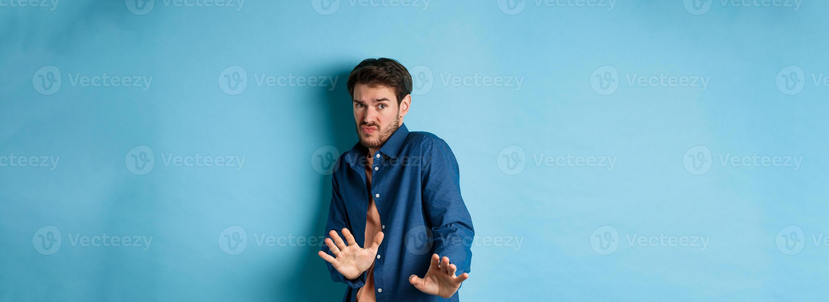 Timid young man asking to keep away, stretch out hands defensive and step back, refusing something disgusting, standing on blue background photo