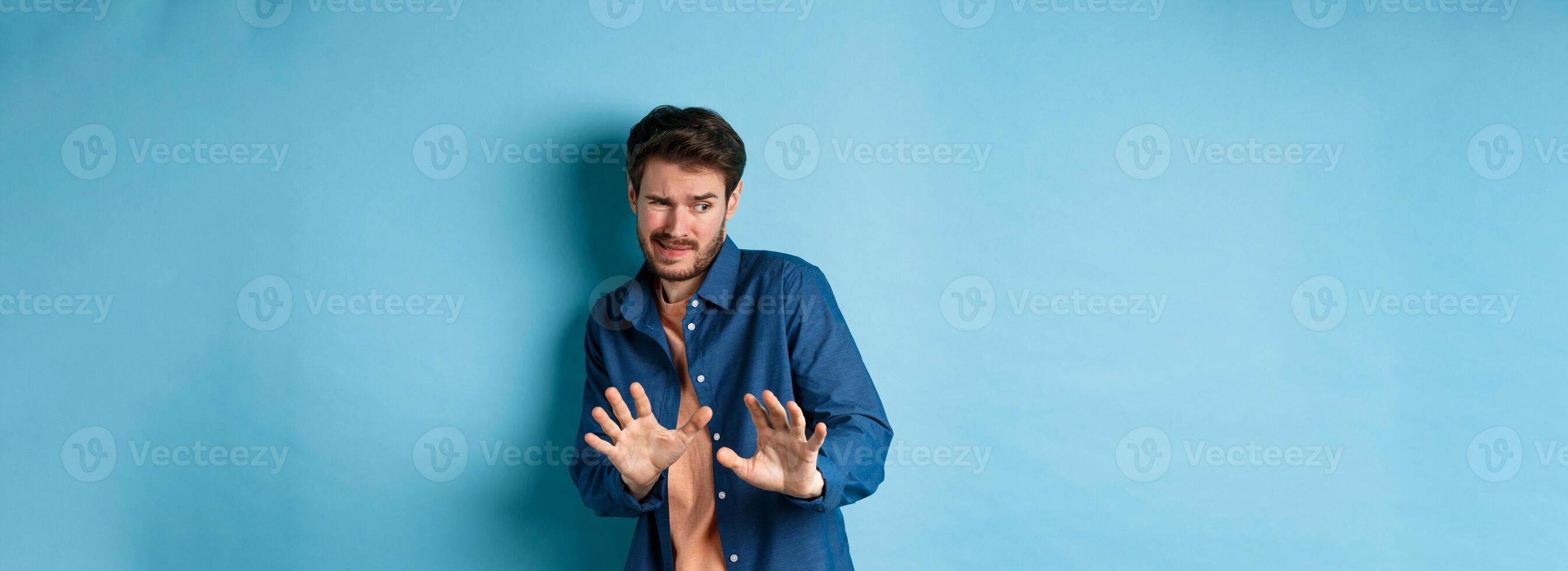 Man cringe and stay away from something nasty, looking aside and stretch out hands to reject bad deal, standing on blue background photo