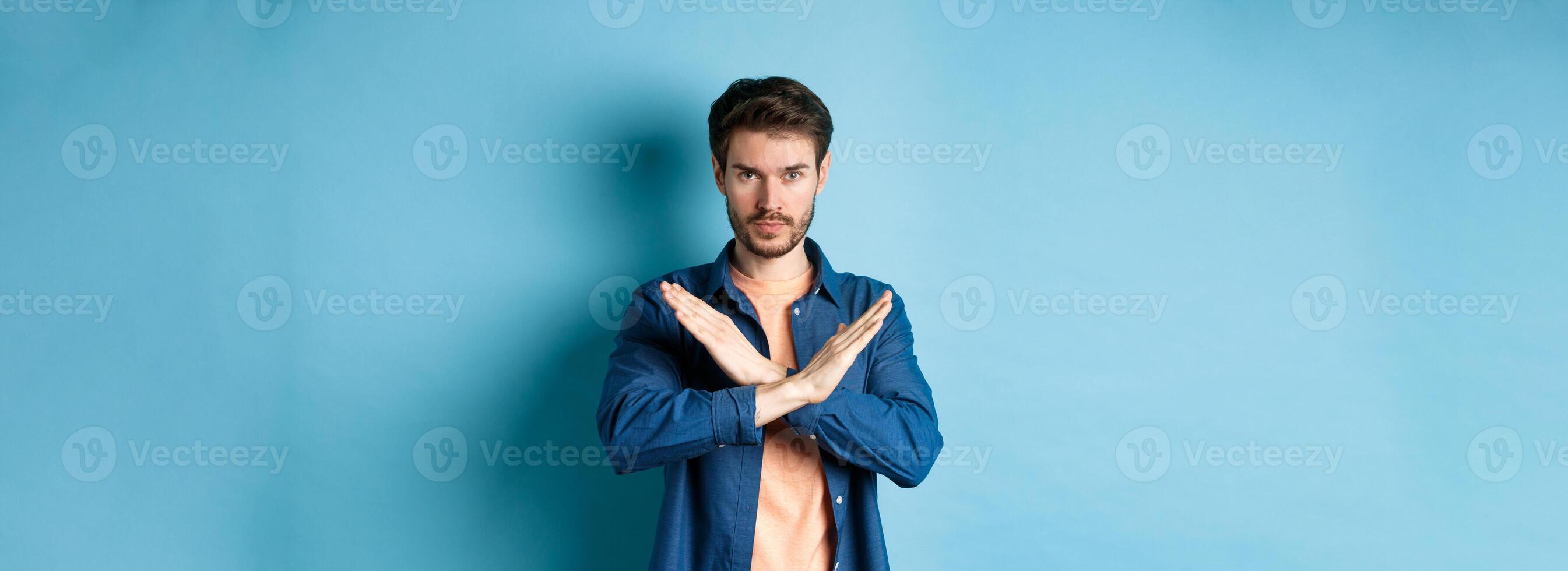 Serious caucasian man tell no, showing cross stop gesture and looking confident at camera, prohibit something bad, standing on blue background photo