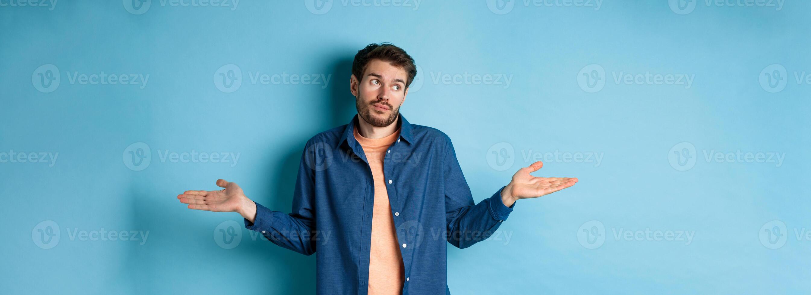 Indecisive young man with beard, look away and shrugging shoulders, know nothing, being clueless, standing on blue background photo