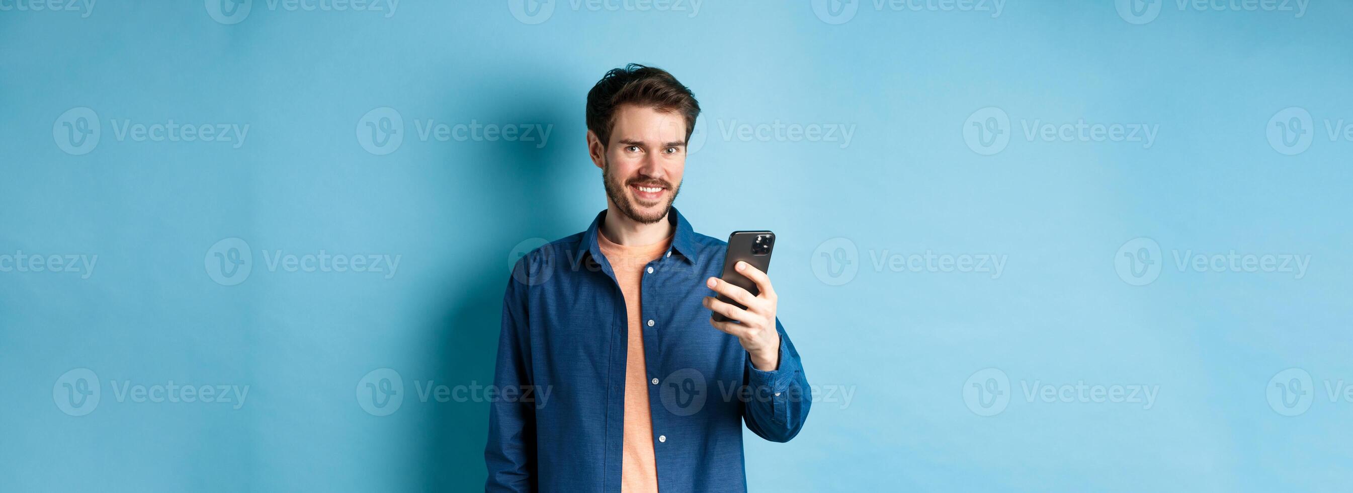 Attractive caucasian man using mobile phone, looking happy and smiling at camera, standing on blue background photo