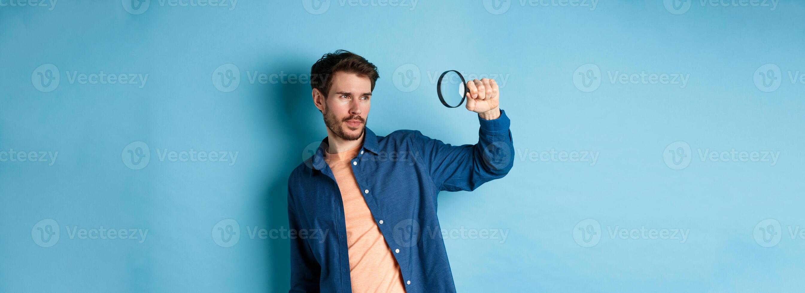 Image of young man searching for something, looking at empty space with magnifying glass, standing on blue background photo
