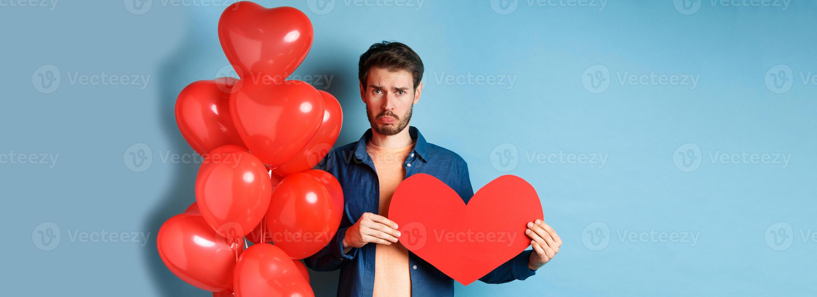 Sad man looking heartbroken and lonely, holding paper red heart and standing near balloons over blue background photo