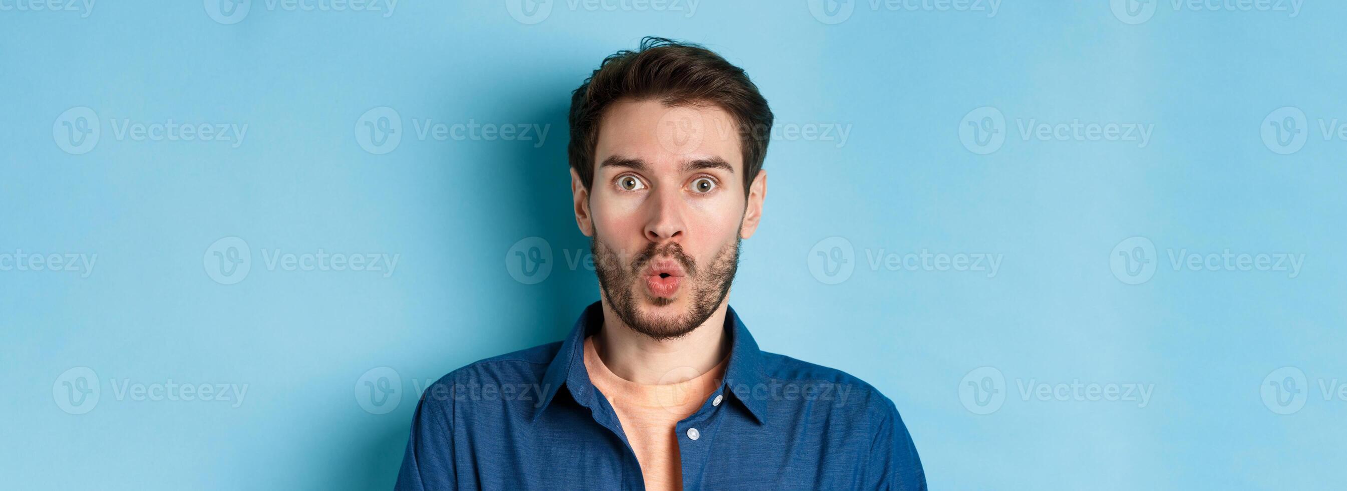 Close up portrait of amazed and surprised man saying wow, staring impressed at camera, checking out cool offer, standing on blue background photo