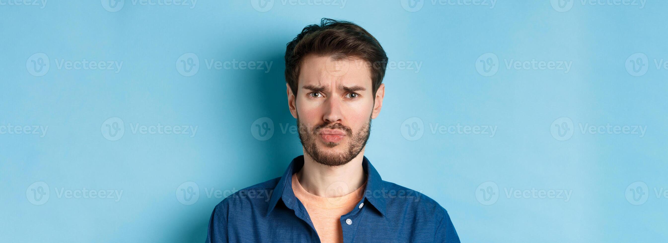 Close up of sad and offended young man sulking at someone, frowning upset, standing on blue background photo