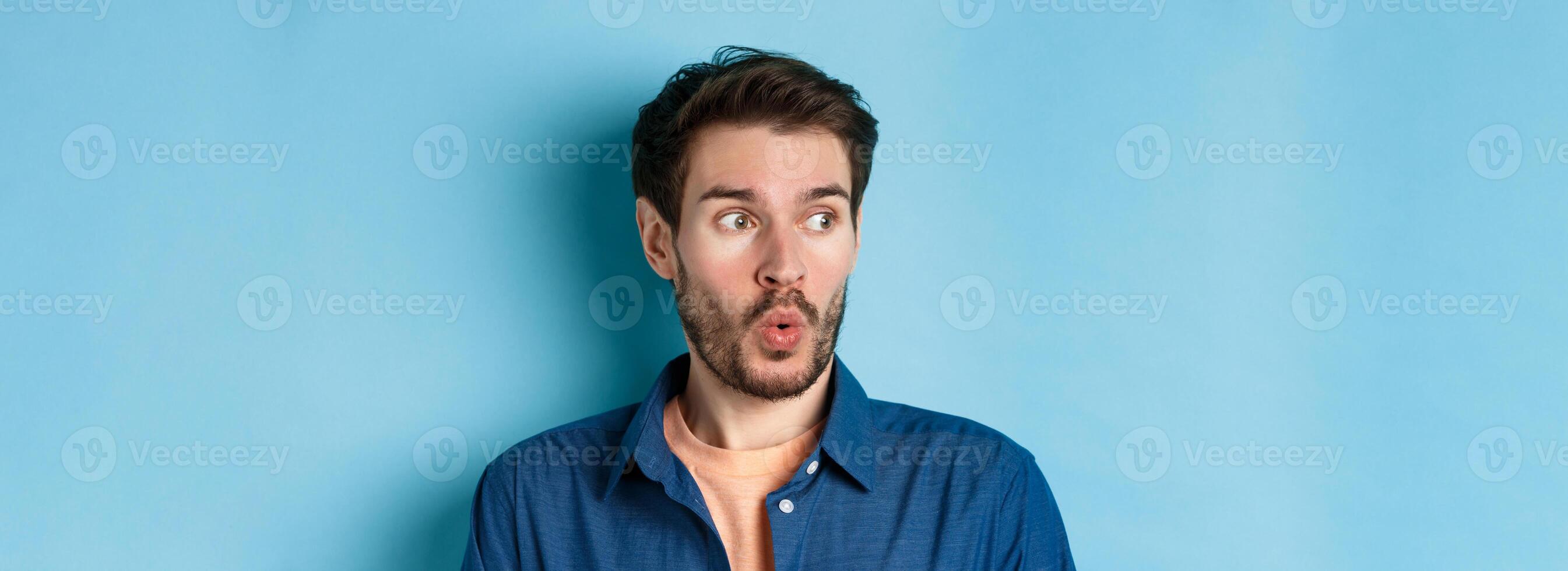 Close up of impressed man say wow and checking out banner, looking left at empty space amazed, standing on blue background photo