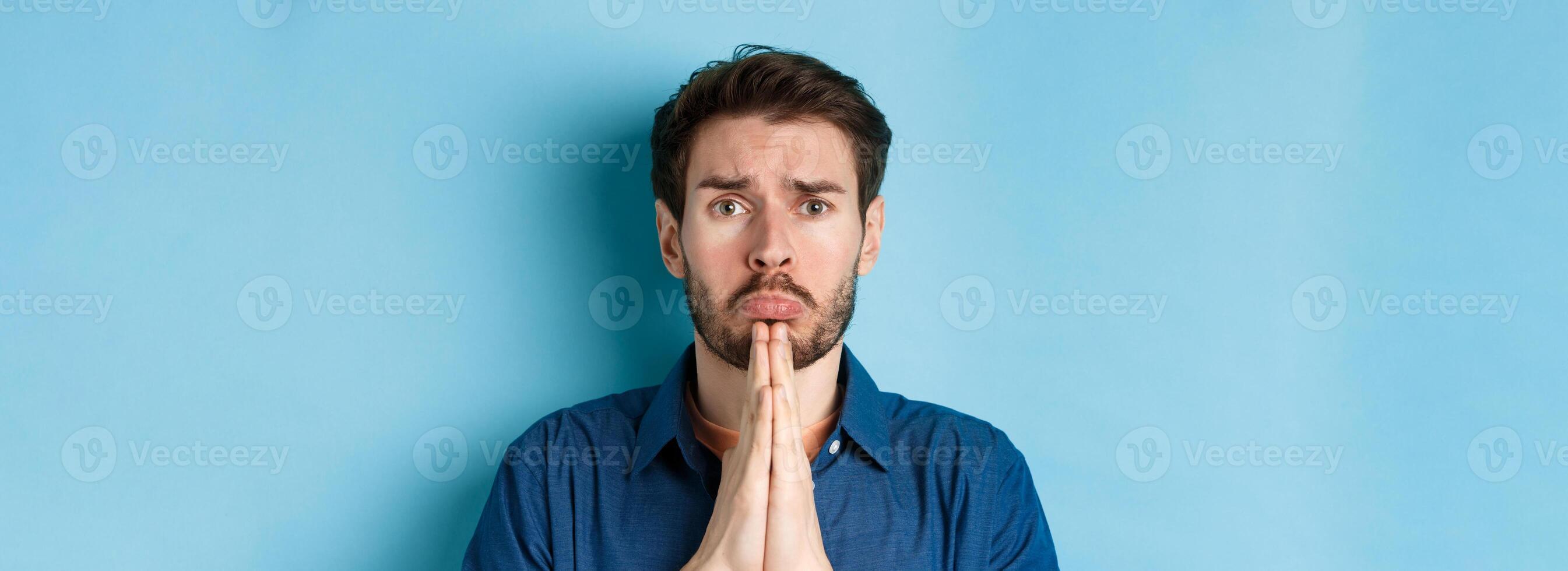 Sad young man begging for help, apoligizing and sobbing miserable, standing on blue background photo