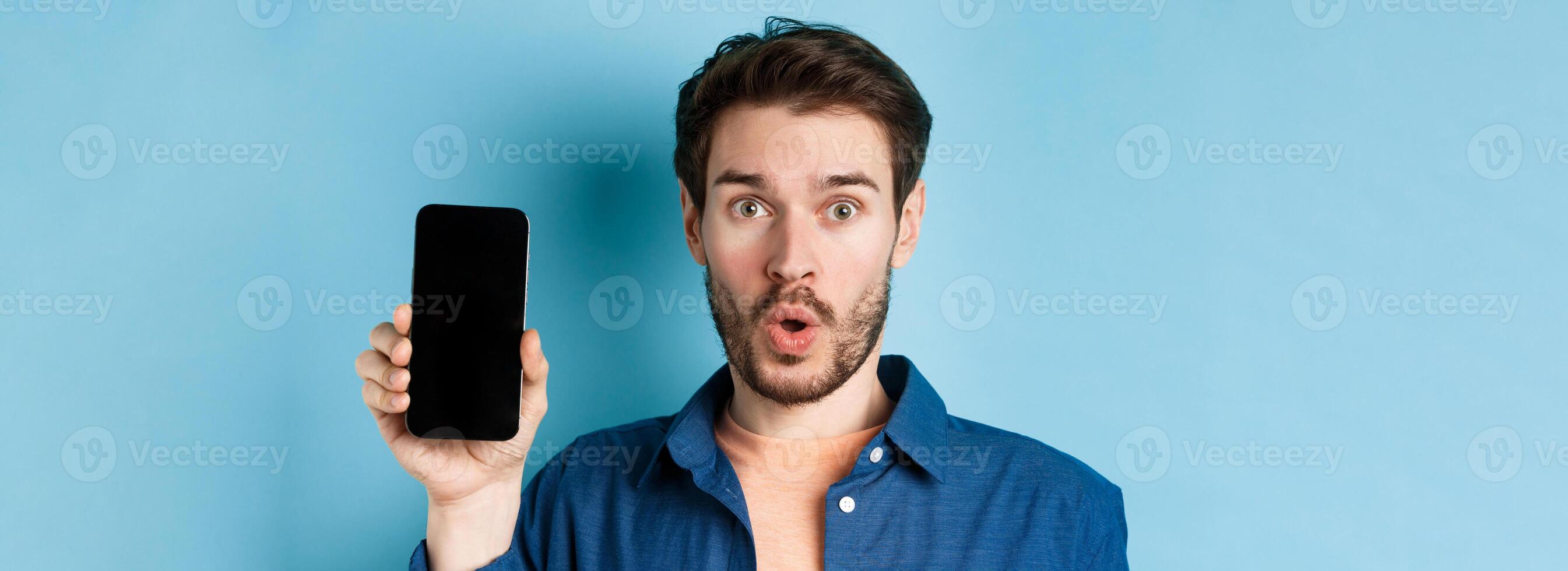 Close up of impressed caucasian man say wow, showing empty smartphone screen, standing on blue background photo