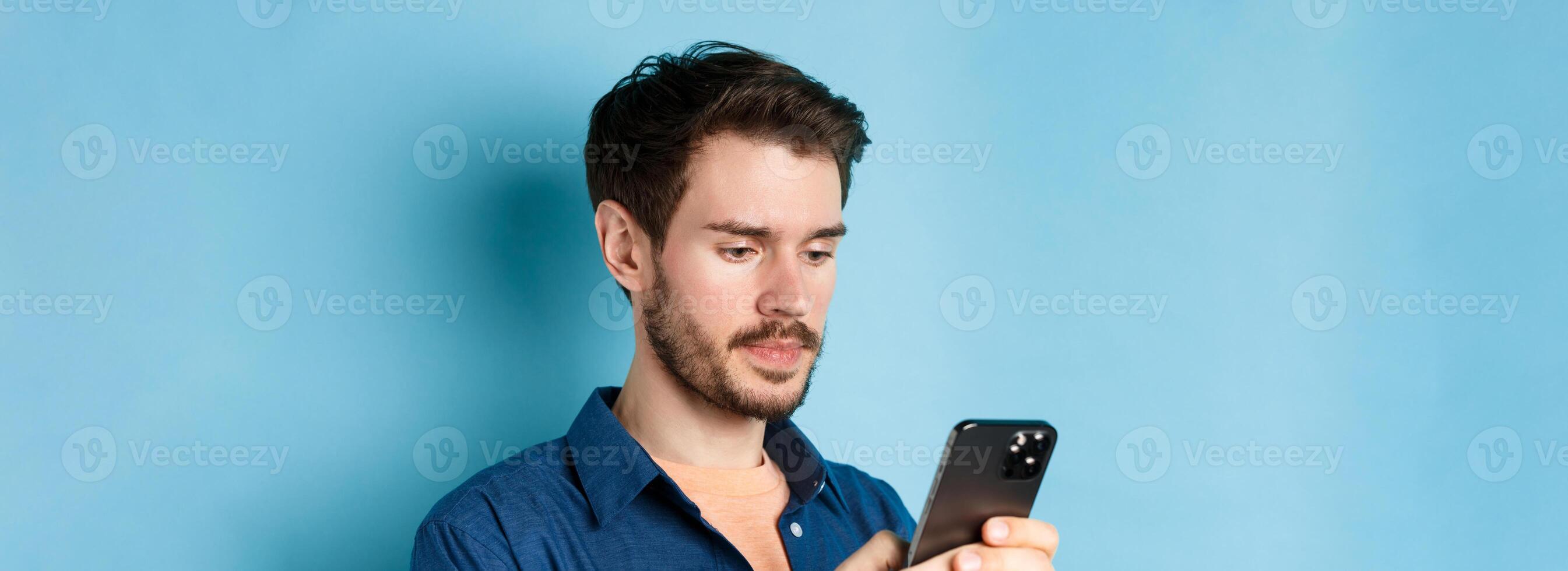 Close up of handsome bearded man reading mobile phone message, looking at screen serious, standing on blue background photo