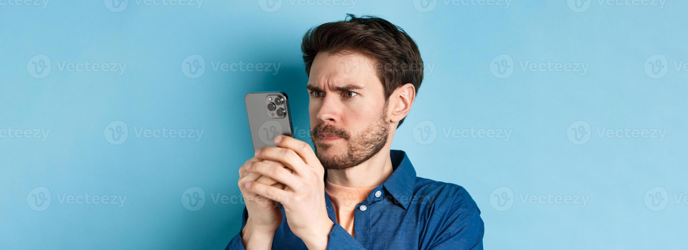 Confused guy staring close at mobile phone screen and frowning, standing on blue background photo