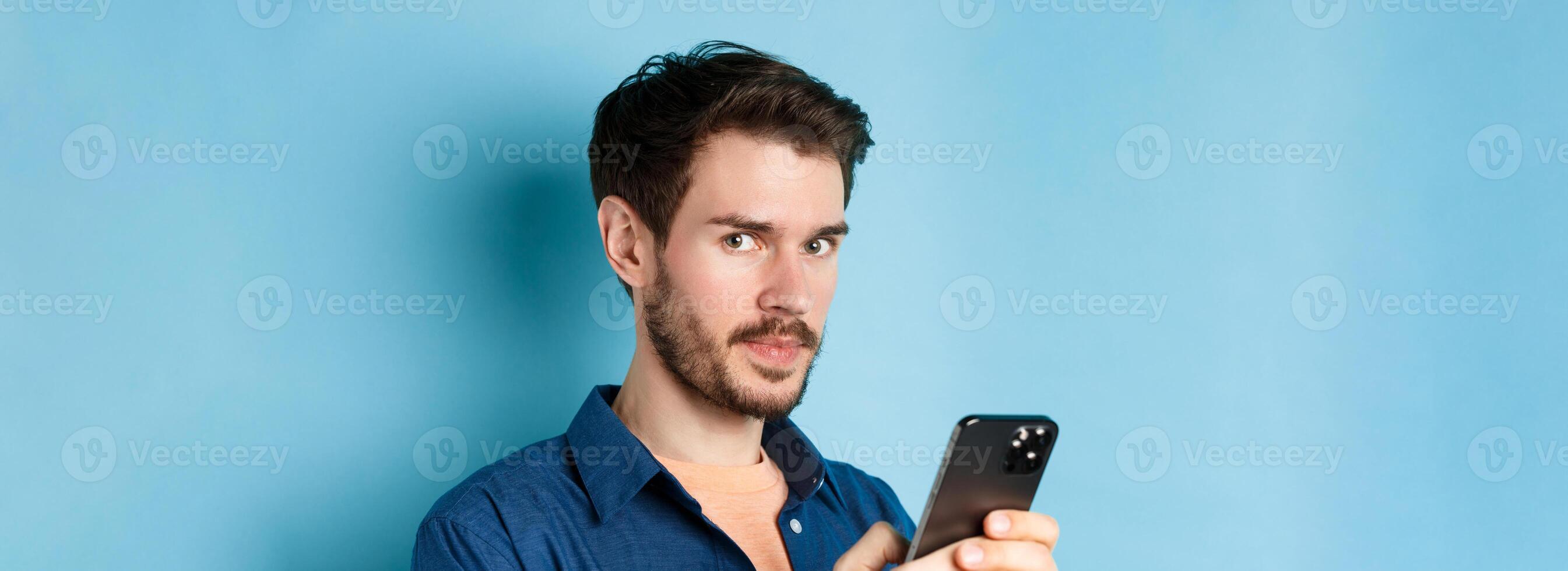 Close-up of handsome european guy using smartphone and looking at camera, standing on blue background photo