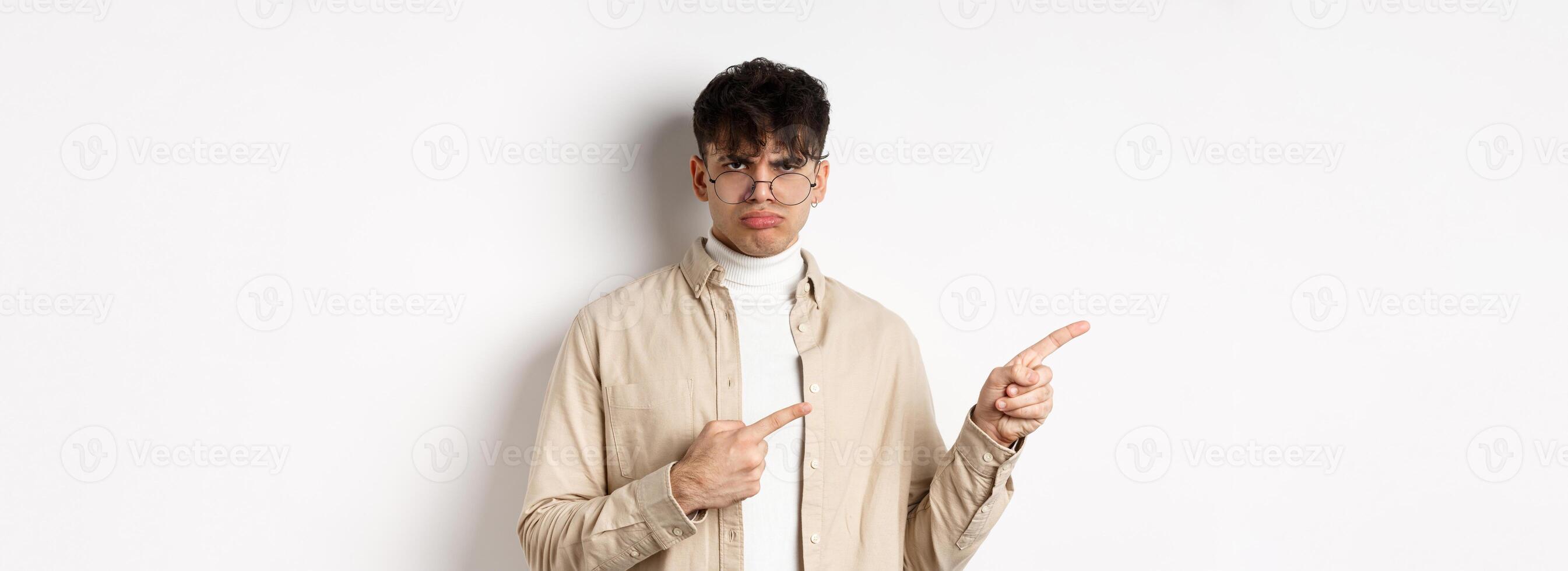 Sad and grumpy boy pointing fingers left, sulking with childish expression, complaining on something bad, standing on white background photo