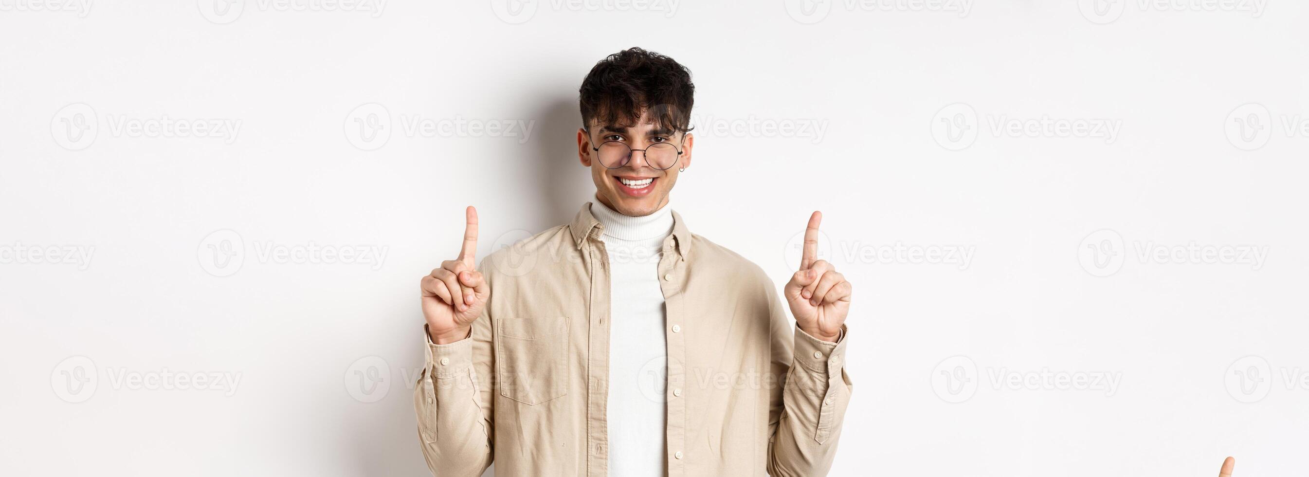 Portrait of handsome hipster guy in glasses showing advertisement and smiling, pointing fingers up at top offer, standing on white background photo