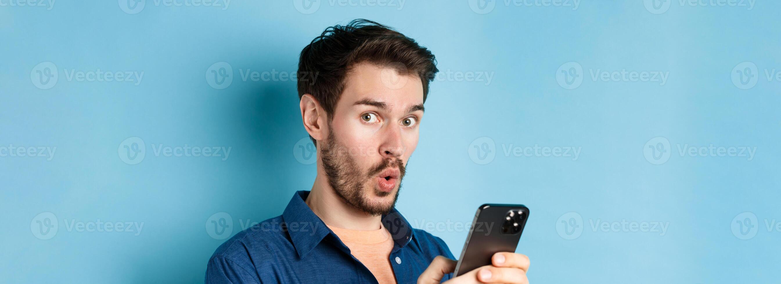 Close up of surprised caucasian man say wow, looking amused at camera, holding smartphone, standing on blue background photo