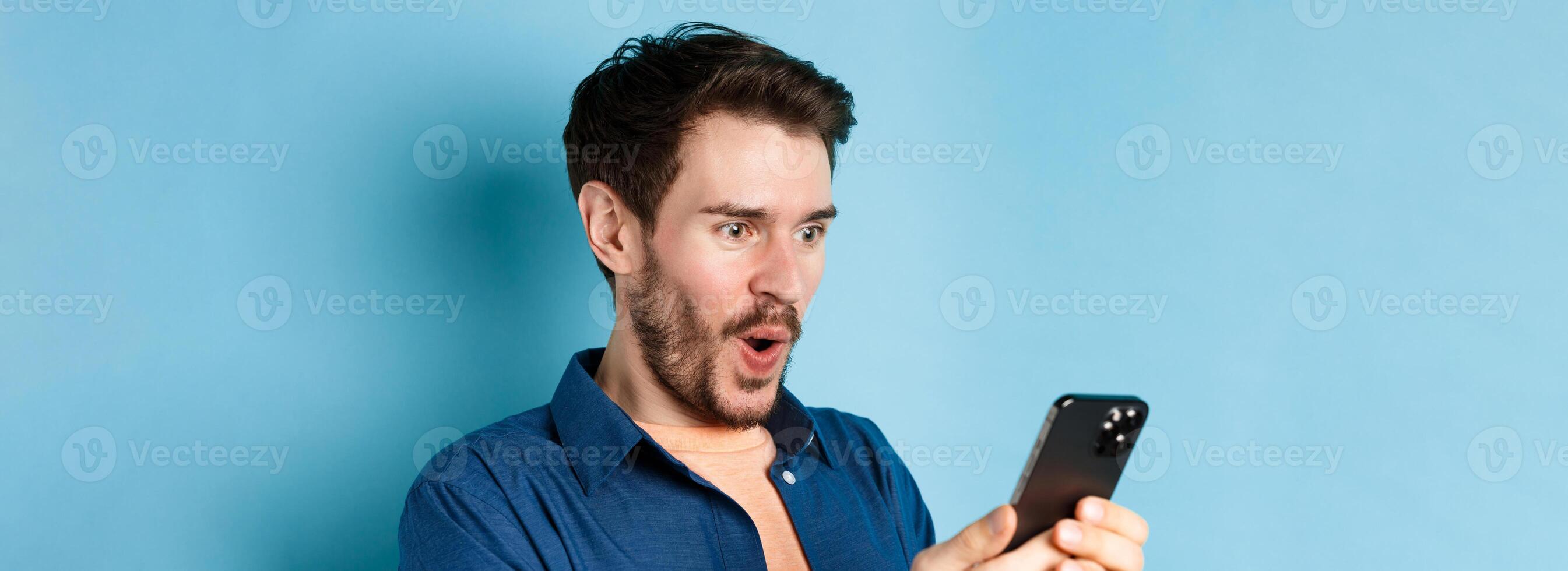 Image of happy young man stare at mobile screen, winning online and cheering, standing on blue background photo