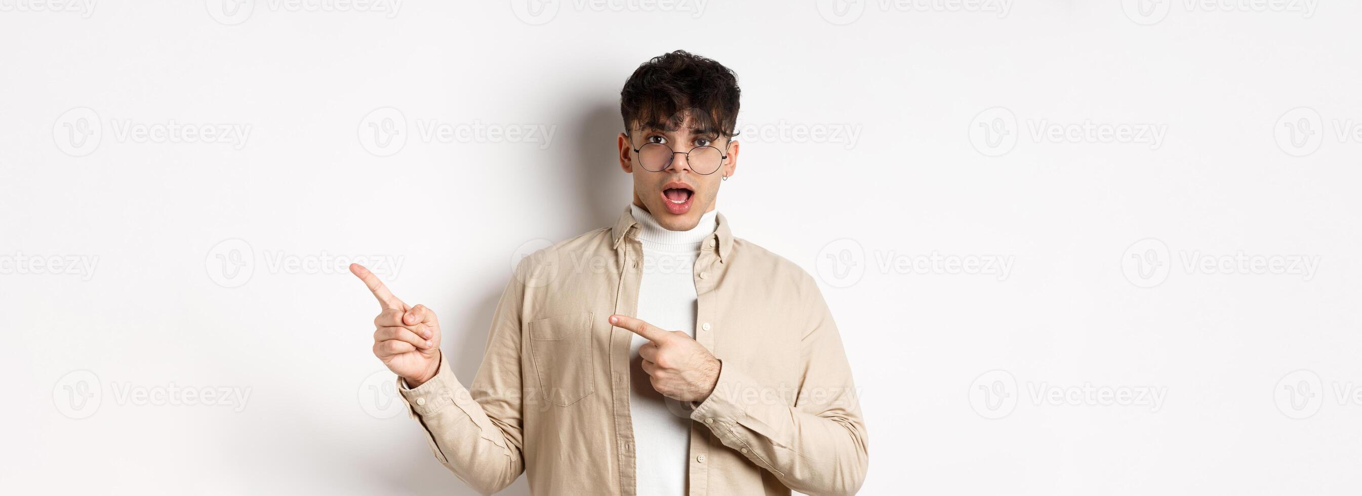 Shocked young man in glasses pointing fingers right at empty space, asking about advertisement, standing on white background photo