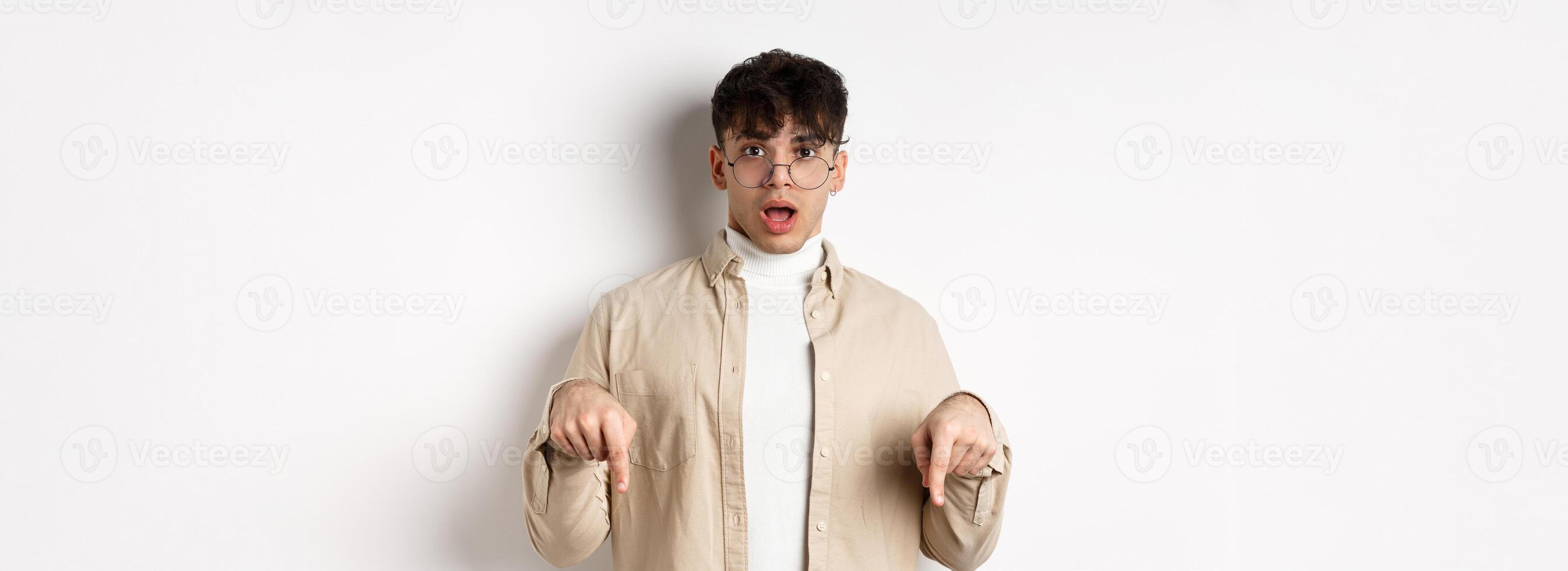 Shocked attractive young man showing something awesome, drop jaw and gasping in awe, pointing fingers down, standing on white background photo