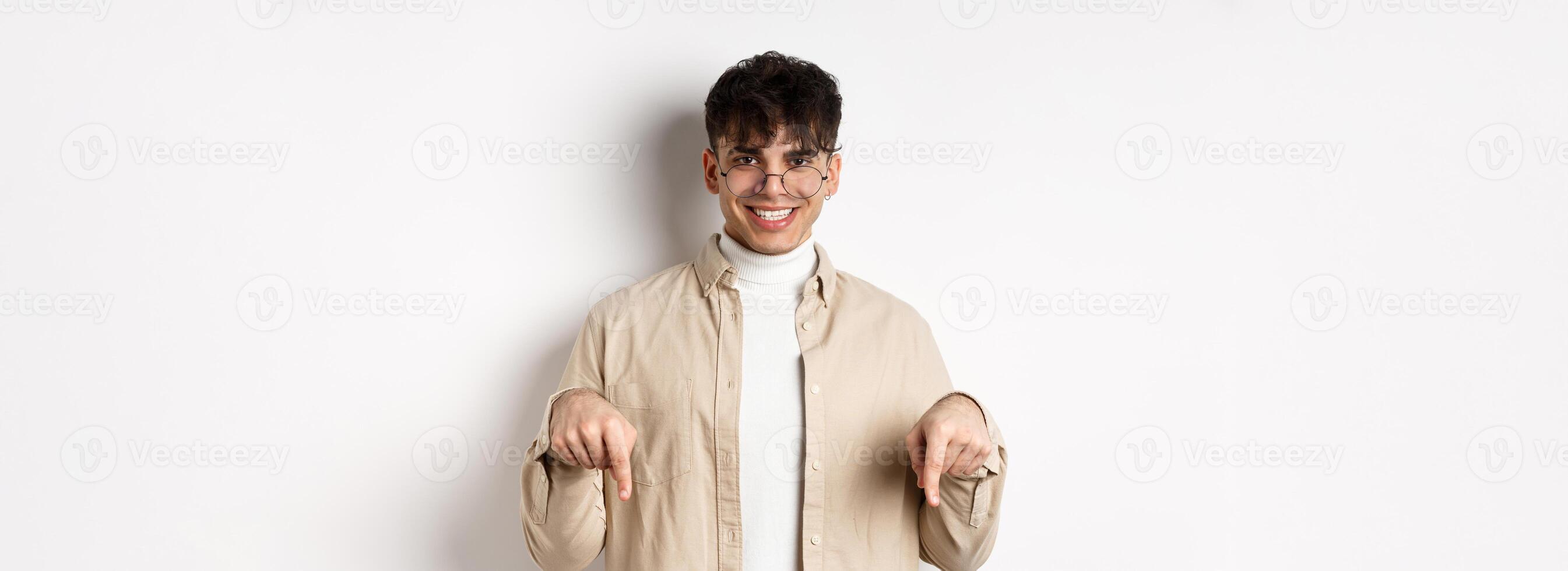 Stylish hipster male model in glasses pointing fingers down, smiling pleased at camera, recommending product, standing on white background photo