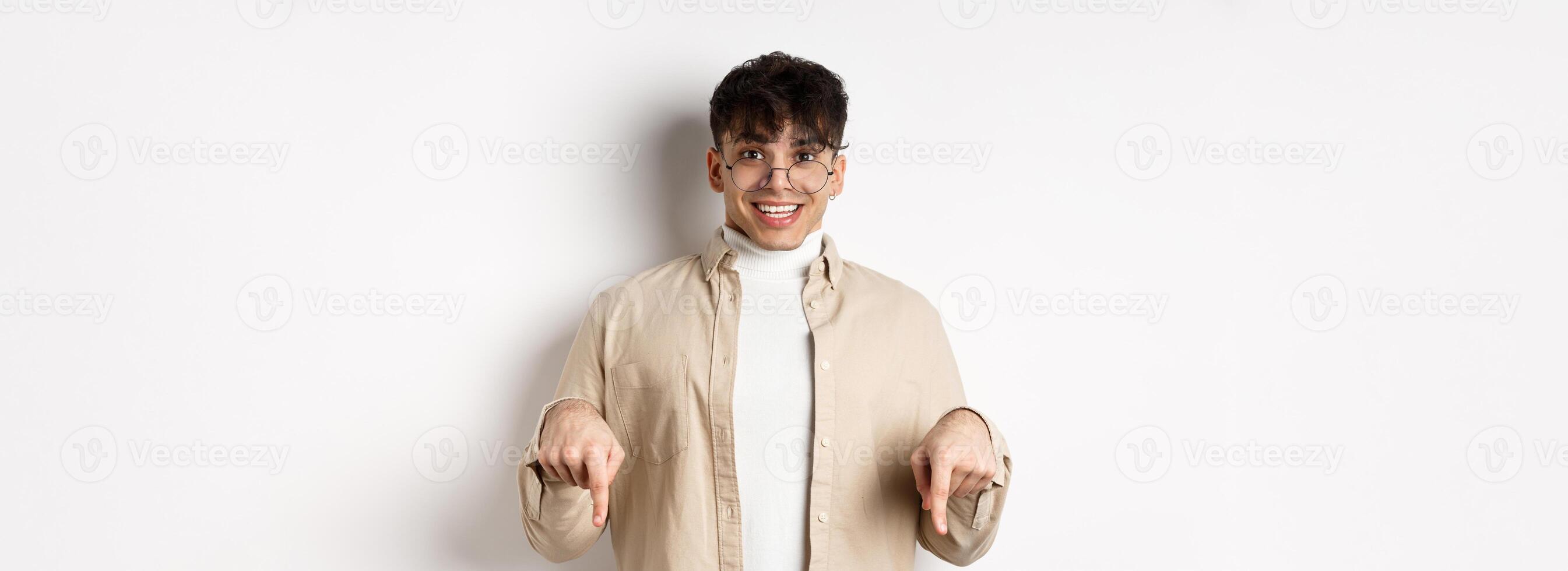 Portrait of happy young guy in glasses showing cool promo, pointing fingers down and smiling excited, standing on white background photo