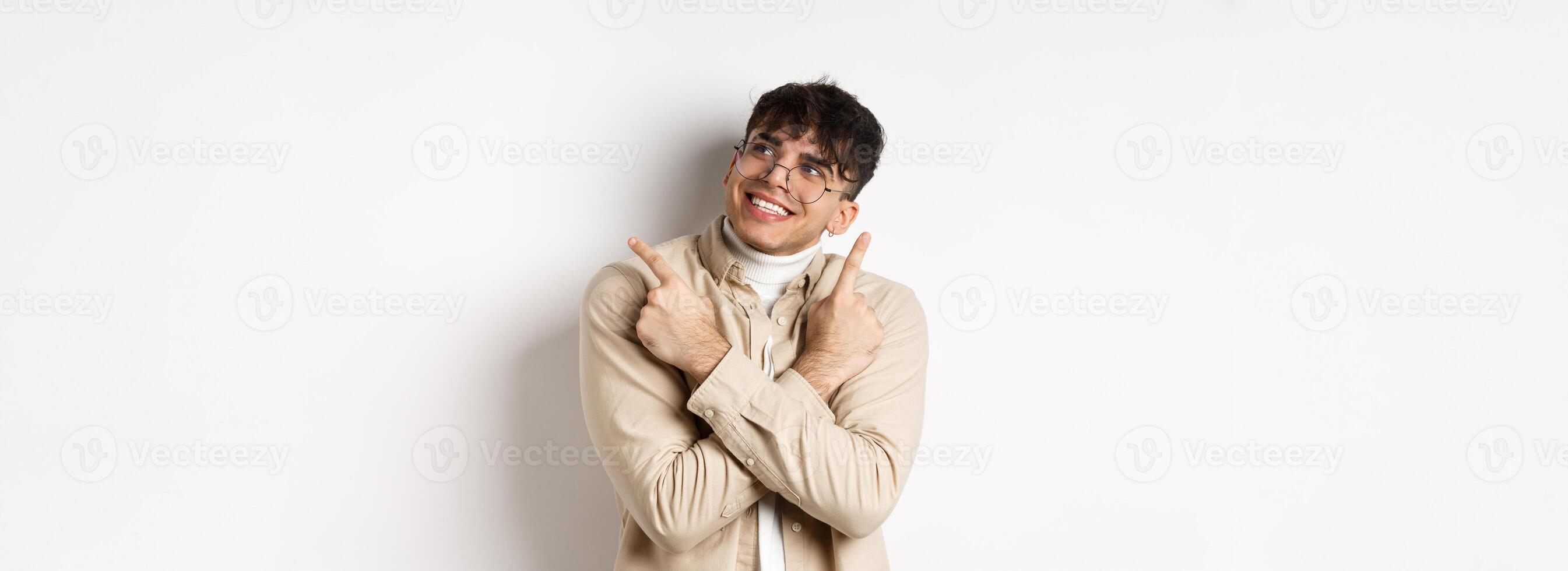 Portrait of handsome smiling guy in glasses pointing fingers sideways, made his choice and look at upper right corner logo, standing on white background photo