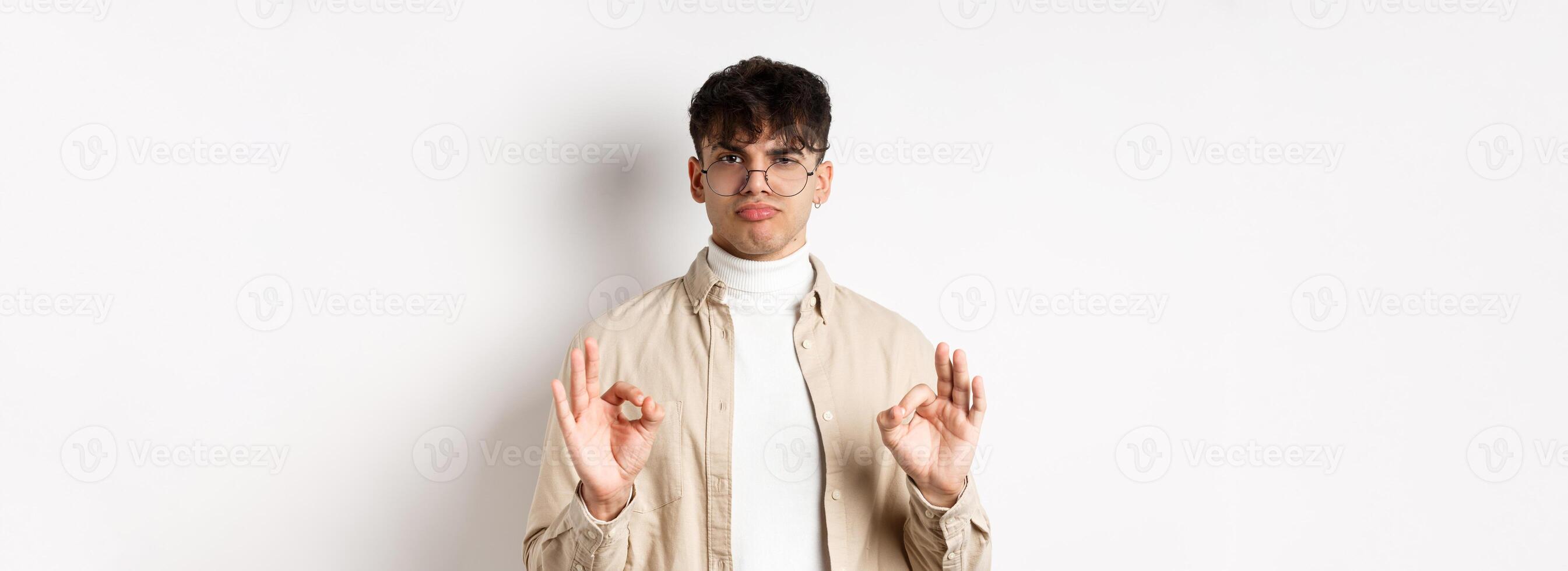 Not bad. Satisfied young man showing okay gestures and nod in approval, praise good work, pleased with results, standing on white background photo