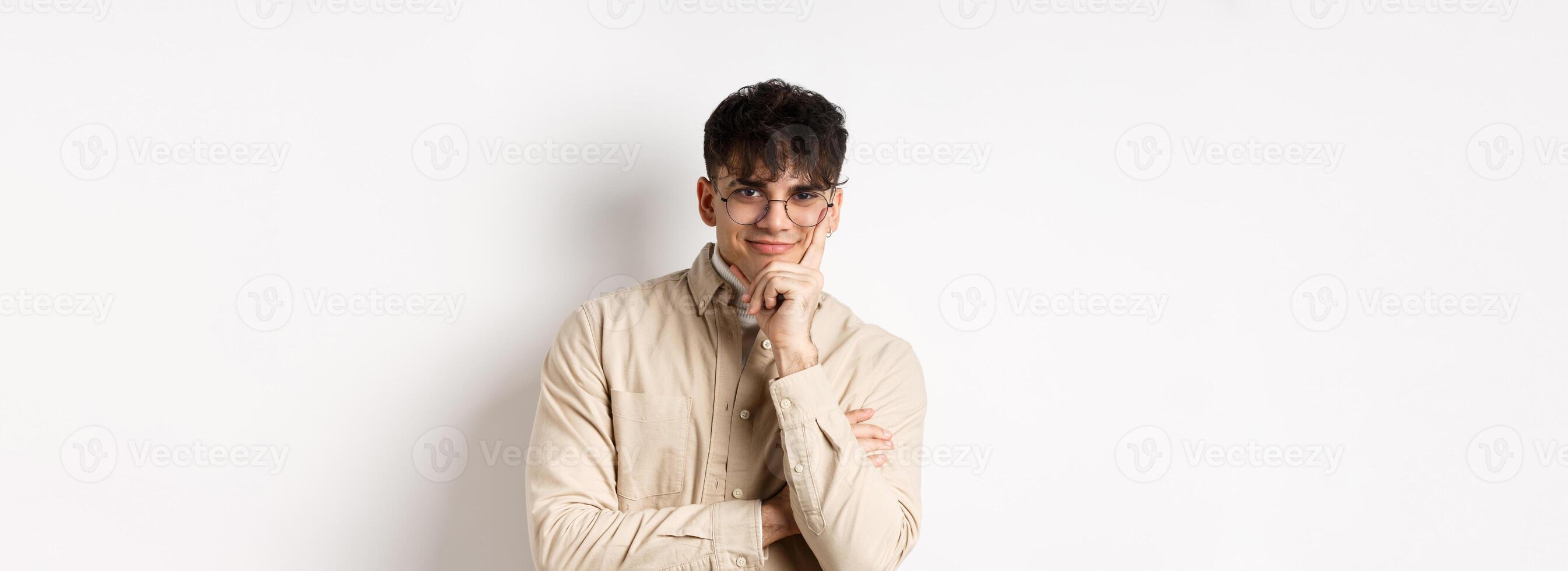 Cunning young man watching something interesting happen, lean face on arm and smiling devious, looking at camera intrigued, standing on white background photo