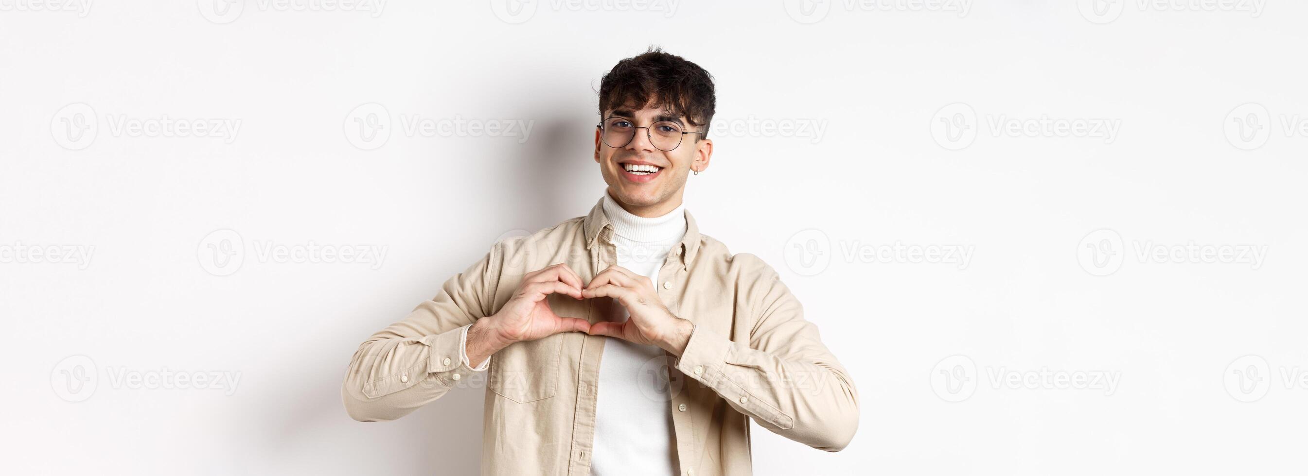 Happy Valentines. Handsome young man in glasses say I love you, showing heart sign and smiling at girlfriend, celebrating lovers day, standing over white background photo
