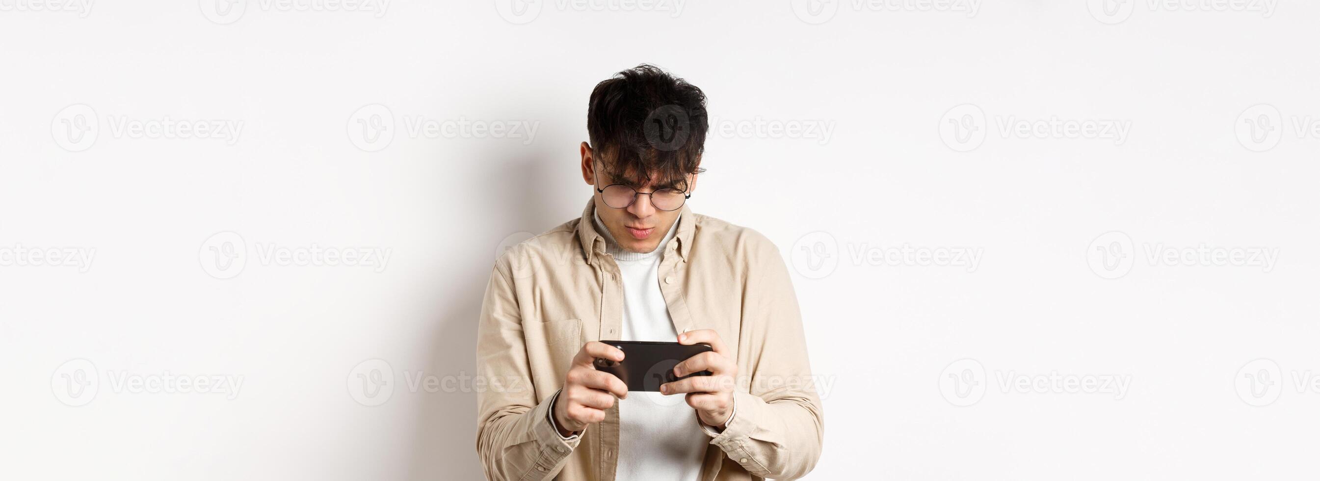 Handsome young man stare at mobile screen and playing game on smartphone, standing on white background photo