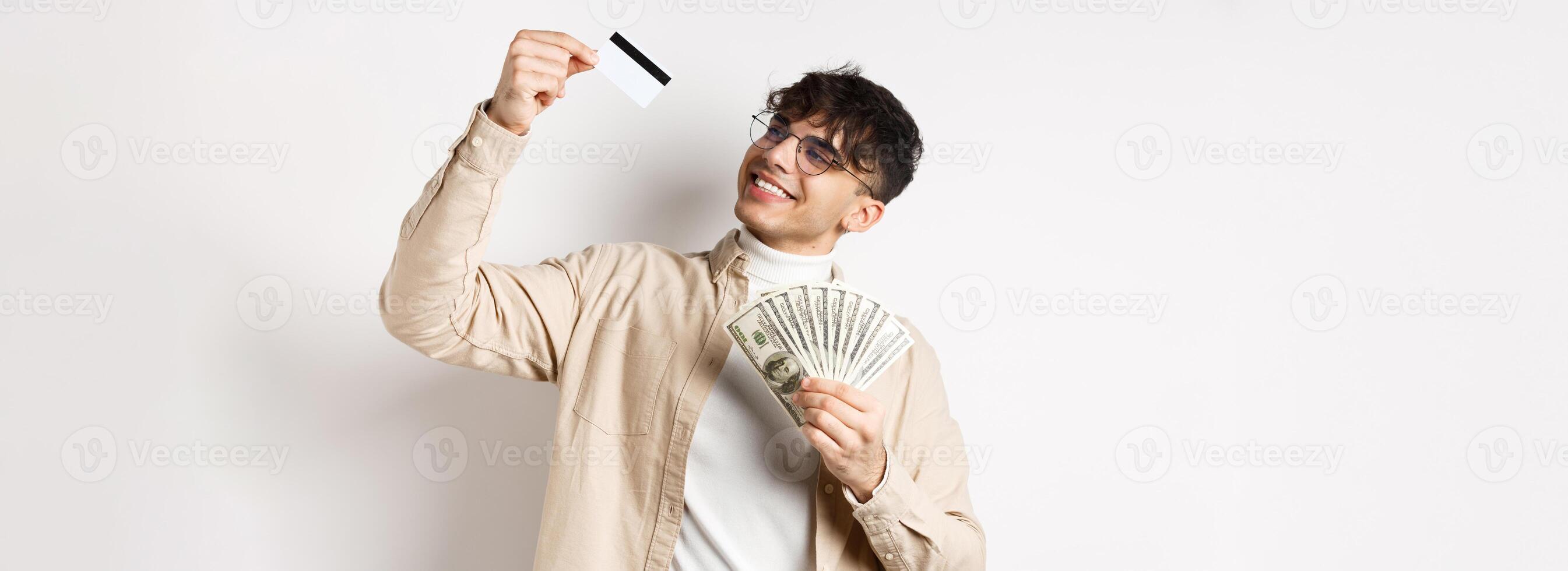 Satisfied young guy holding money, looking pleased and smiling at plastic credit card, choose contactless payment, standing on white background photo