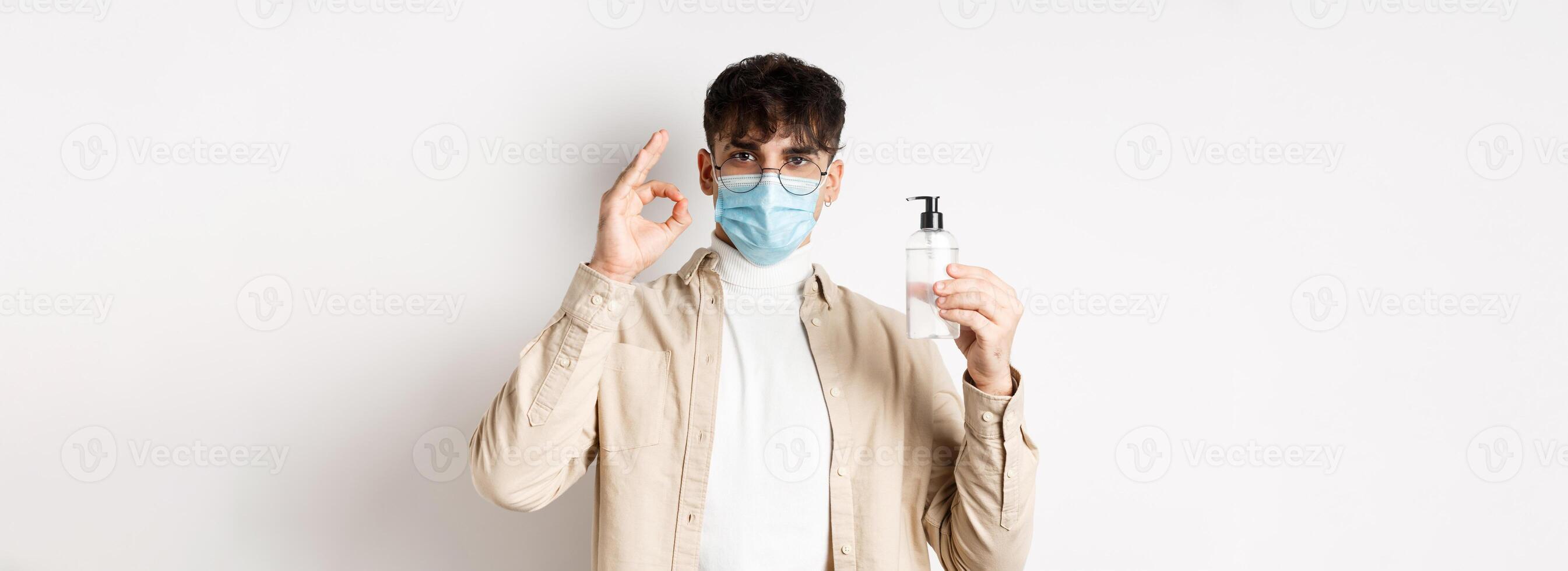 Health, covid and quarantine concept. Portrait of natural guy in glasses and face mask showing bottle of good hand sanitizer, make okay sign, recommending product, white background photo
