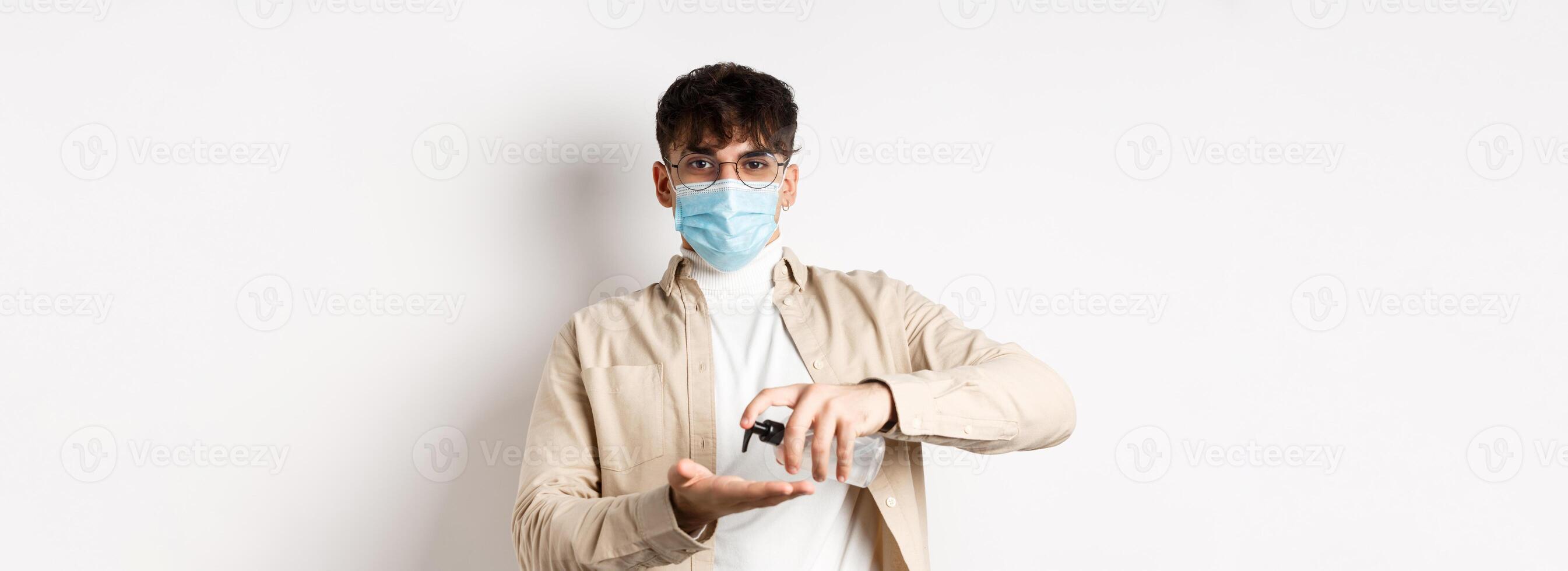 Health, covid and quarantine concept. Young hispanic guy in glasses and face mask using hand sanitizer, apply antiseptic and look at camera, standing on white background photo