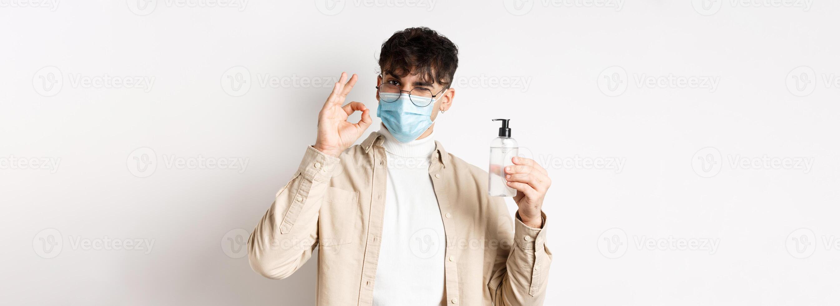 Health, covid and quarantine concept. Portrait of natural guy in glasses and face mask showing bottle of good hand sanitizer, make okay sign, recommending product, white background photo