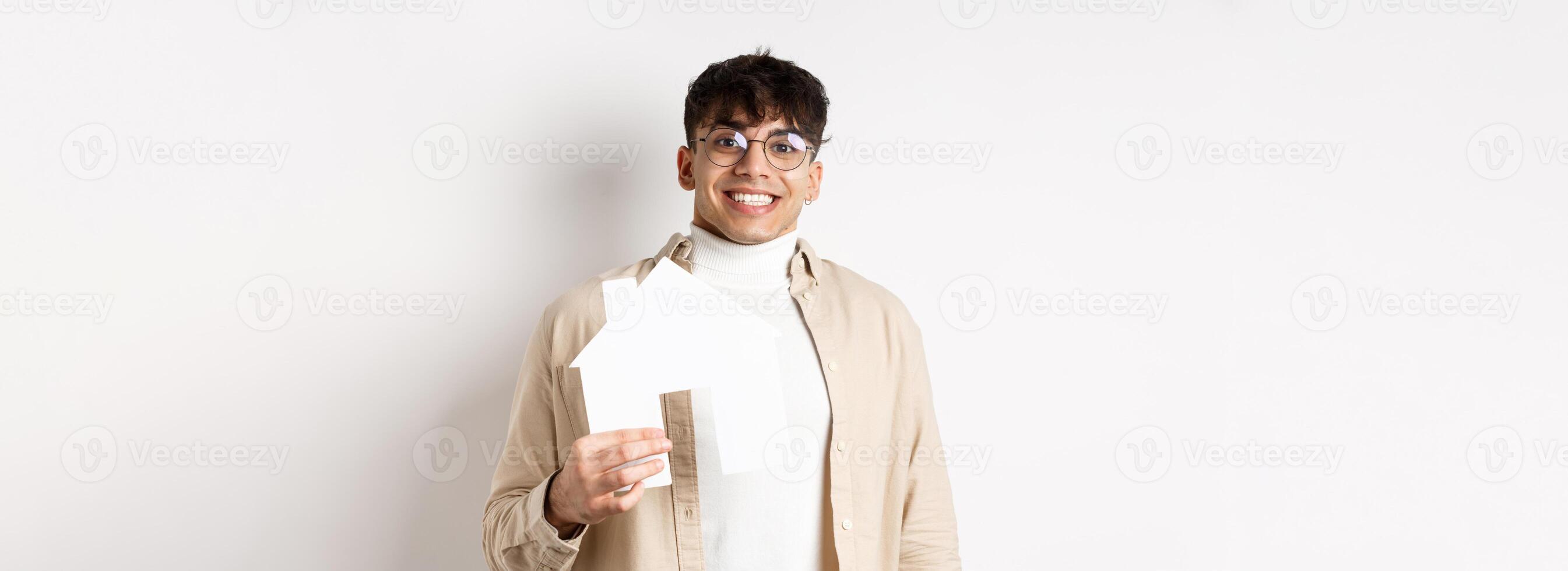 Real estate and mortgage concept. Cheerful young guy renting apartment, showing paper house cutout and smiling happy, buying property, white background photo