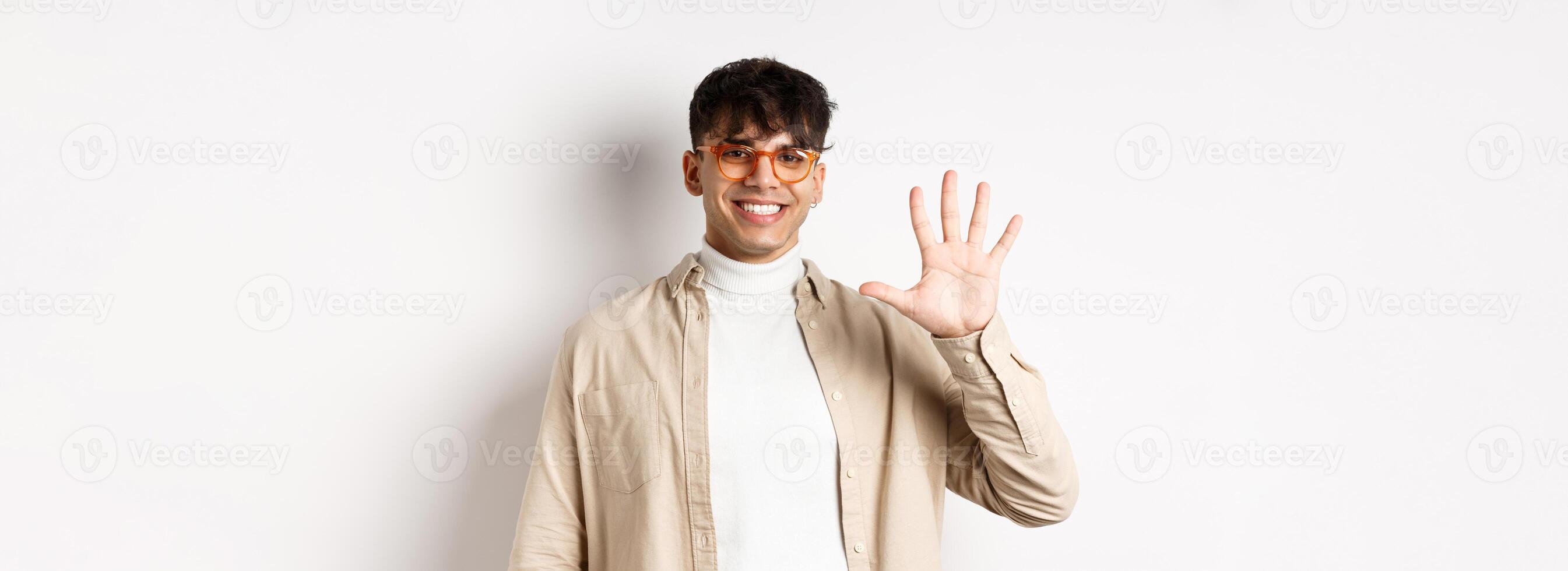 Modern guy in glasses and stylish outfit, showing five fingers number and smiling, making order, standing on white background photo