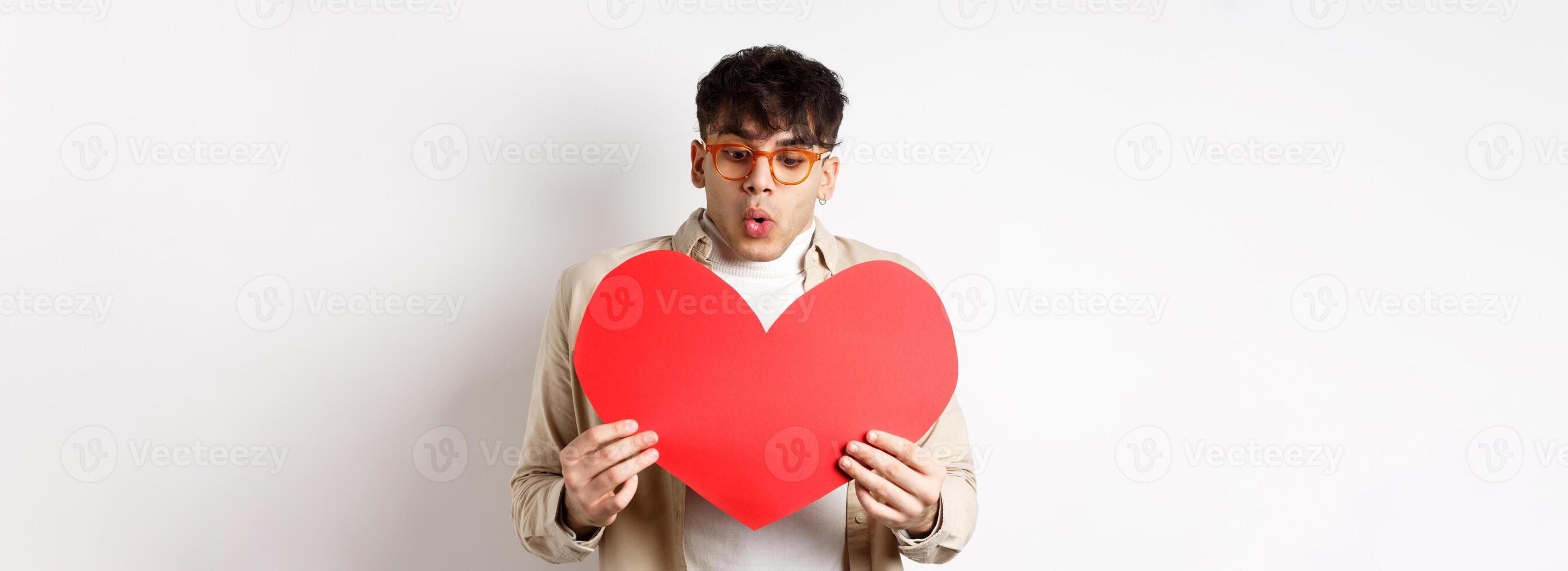 Surprised handsome man receive big red heart postcard on Valentines day, looking at gift with amazement, enjoying lovers day, standing over white background photo