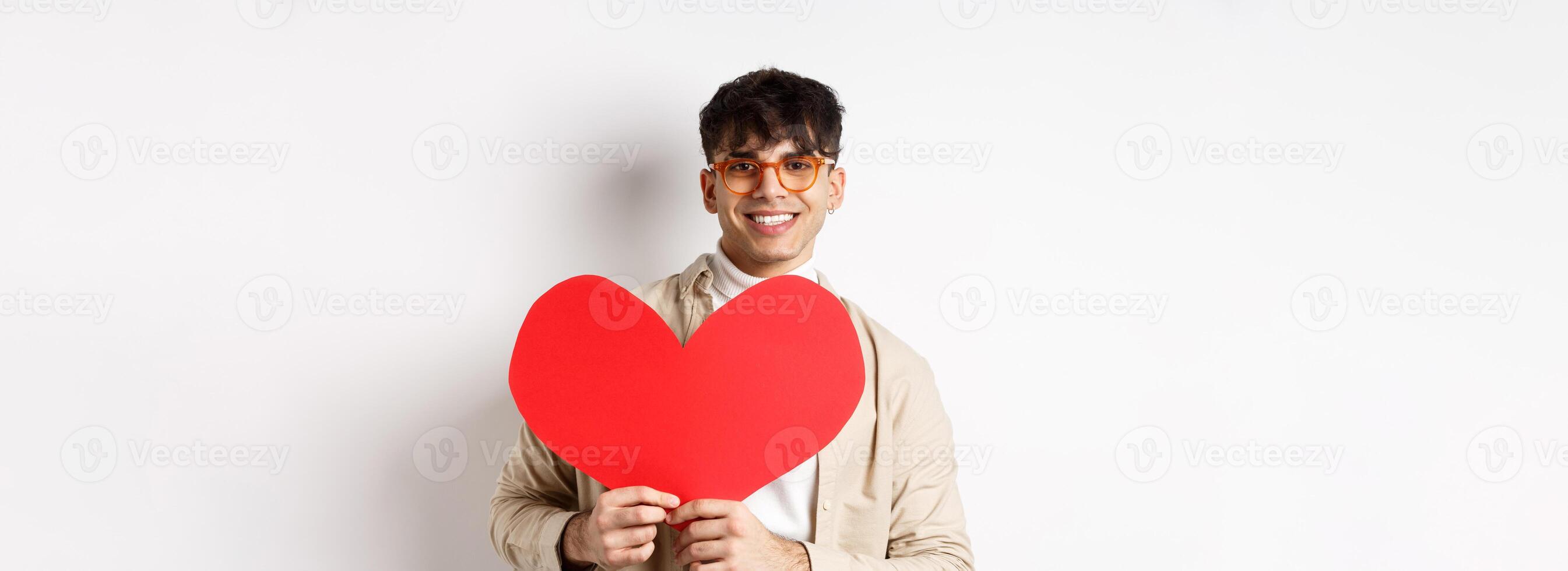 Handsome hipster guy with sunglasses and earring, waiting for true love on Valentines day, holding big red heart and smiling, standing over white background photo