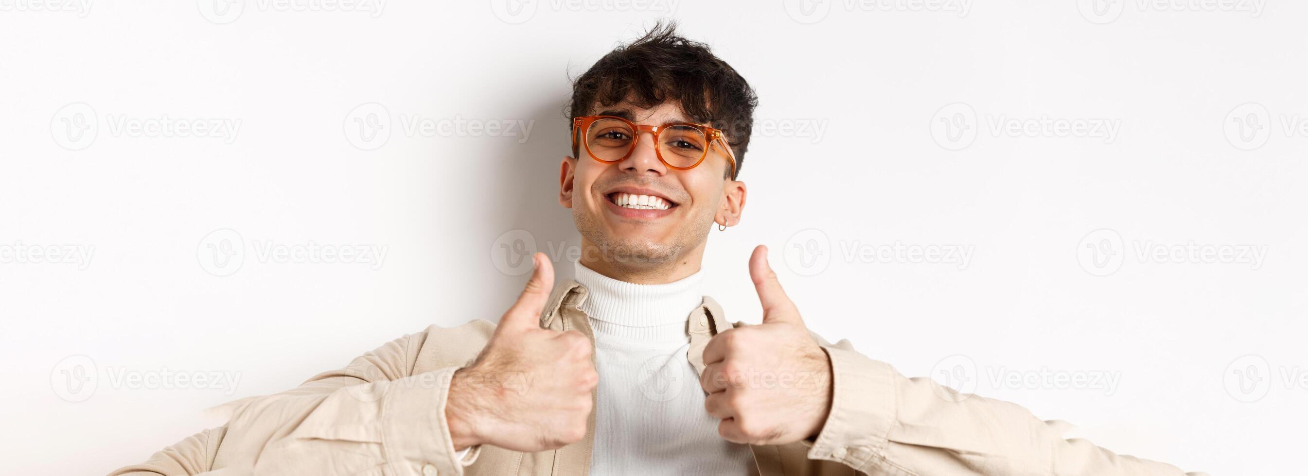 Close-up of satisfied happy guy showing thumbs up and smiling with white teeth, wearing eyewear, standing on white background photo