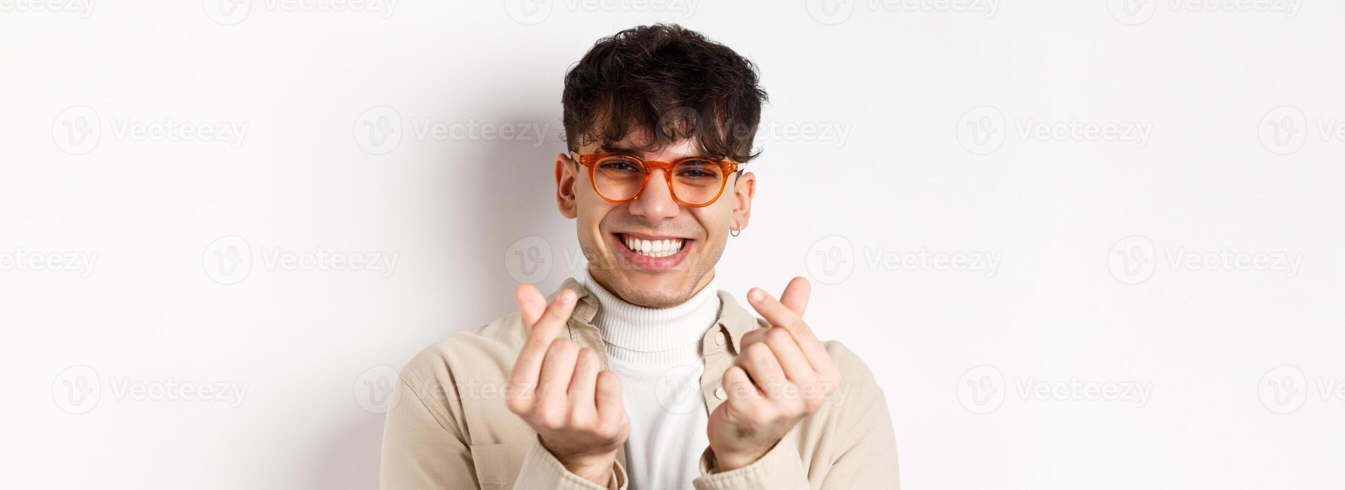 Cute young guy in glasses smiling and showing finger hearts, standing on white background photo