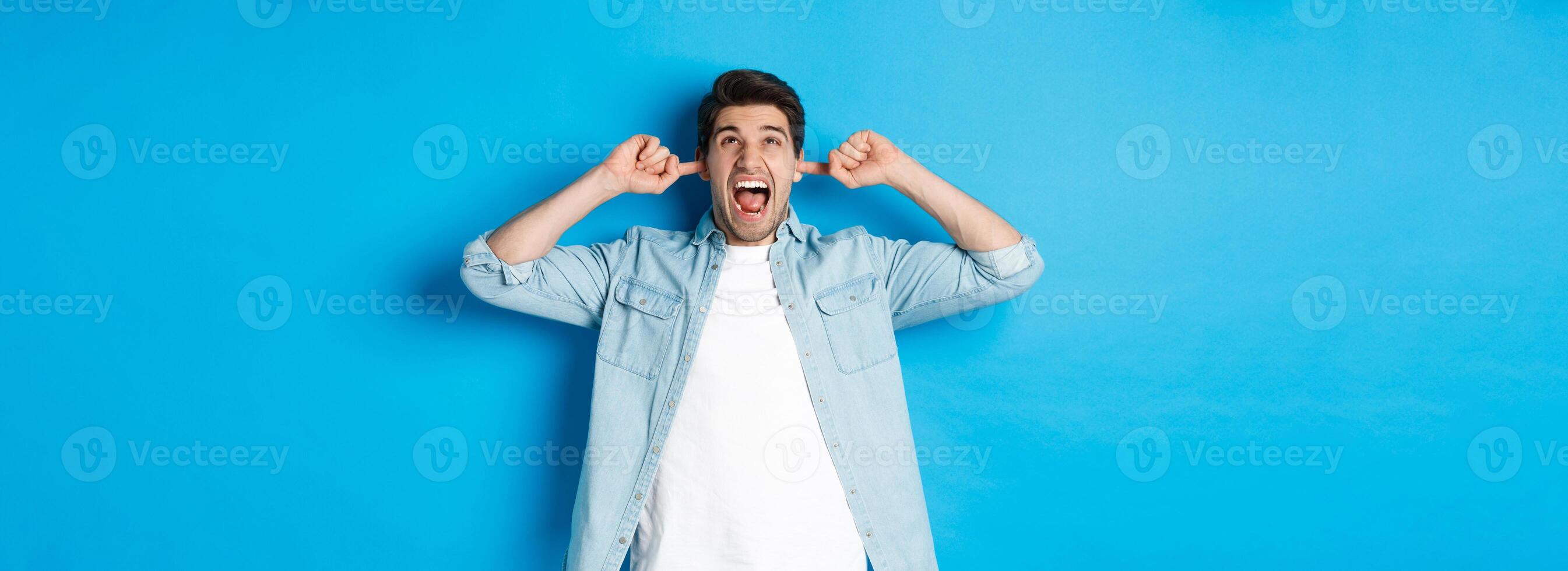 Irritated man close ears from noisy neighbours, looking up and shouting annoyed, hearing loud music, standing over blue background photo