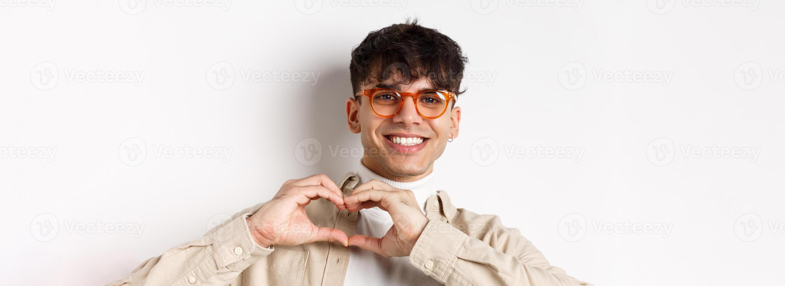 Close-up of young natural guy in glasses smiling, showing heart love you gesture, standing on white background photo