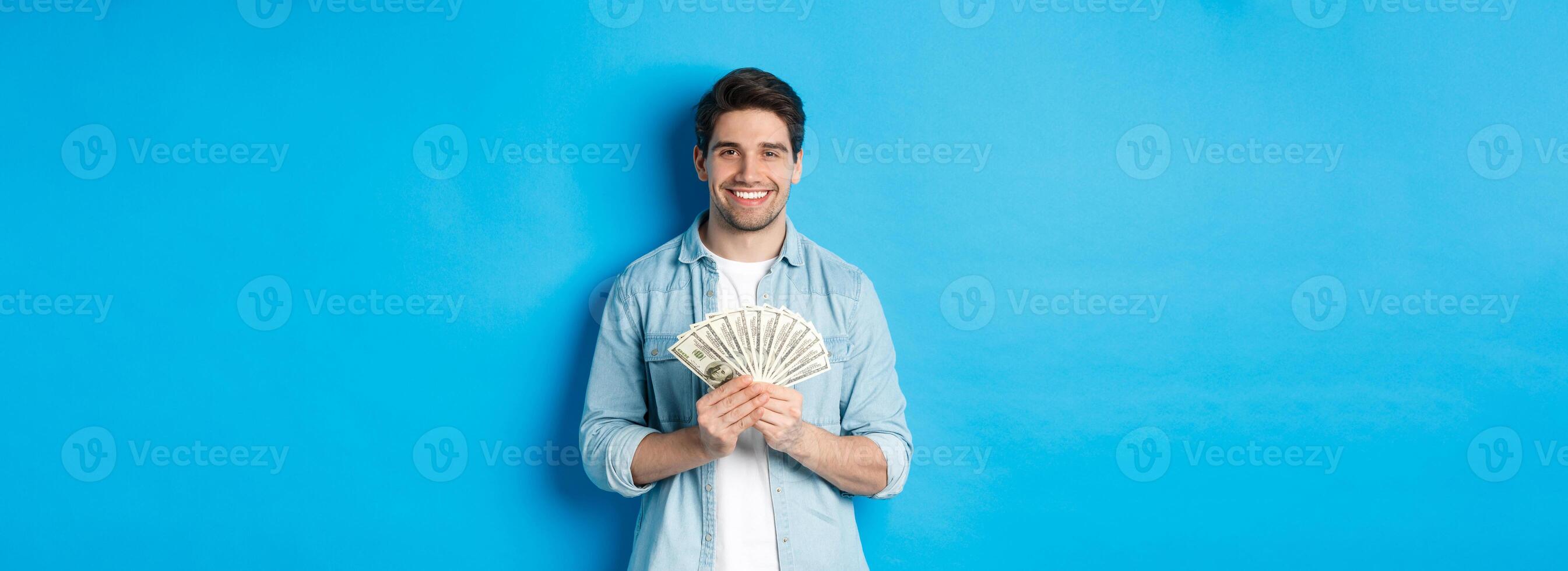 Handsome smiling man holding money, concept of finance and banking, standing over blue background photo