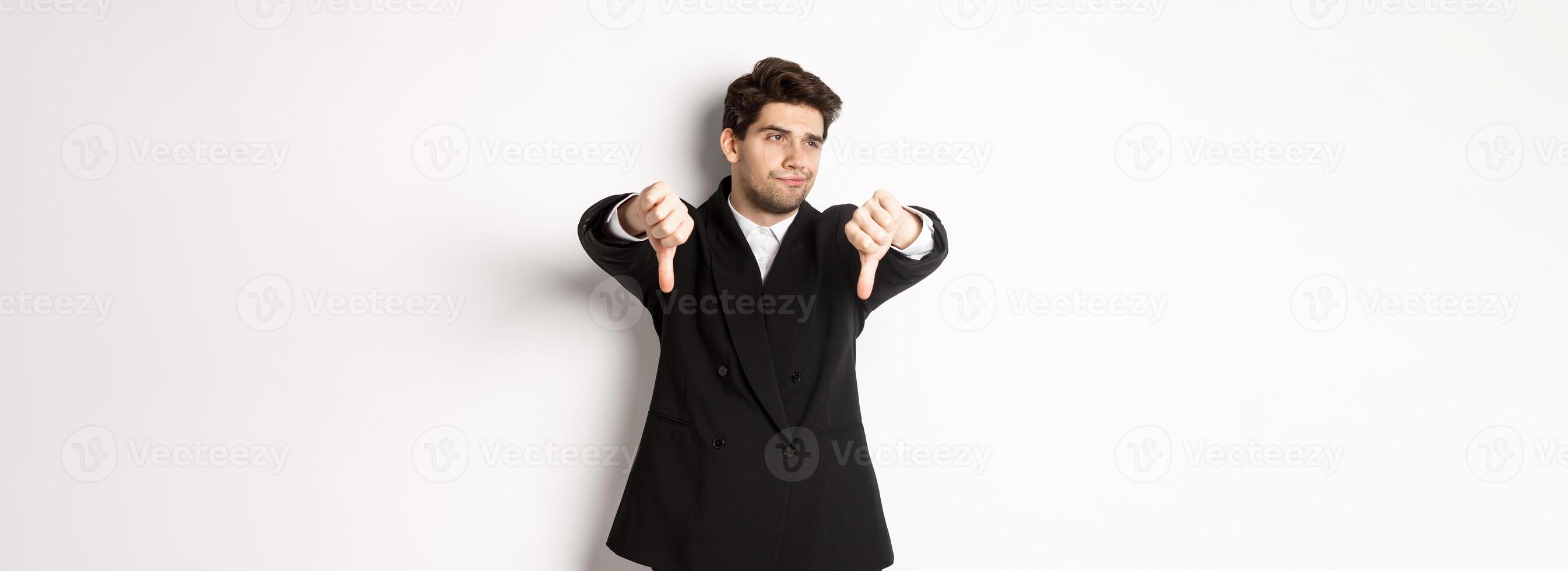 Skeptical and displeased man in black suit, looking away reluctant, showing thumbs-down, dislike something, standing over white background photo