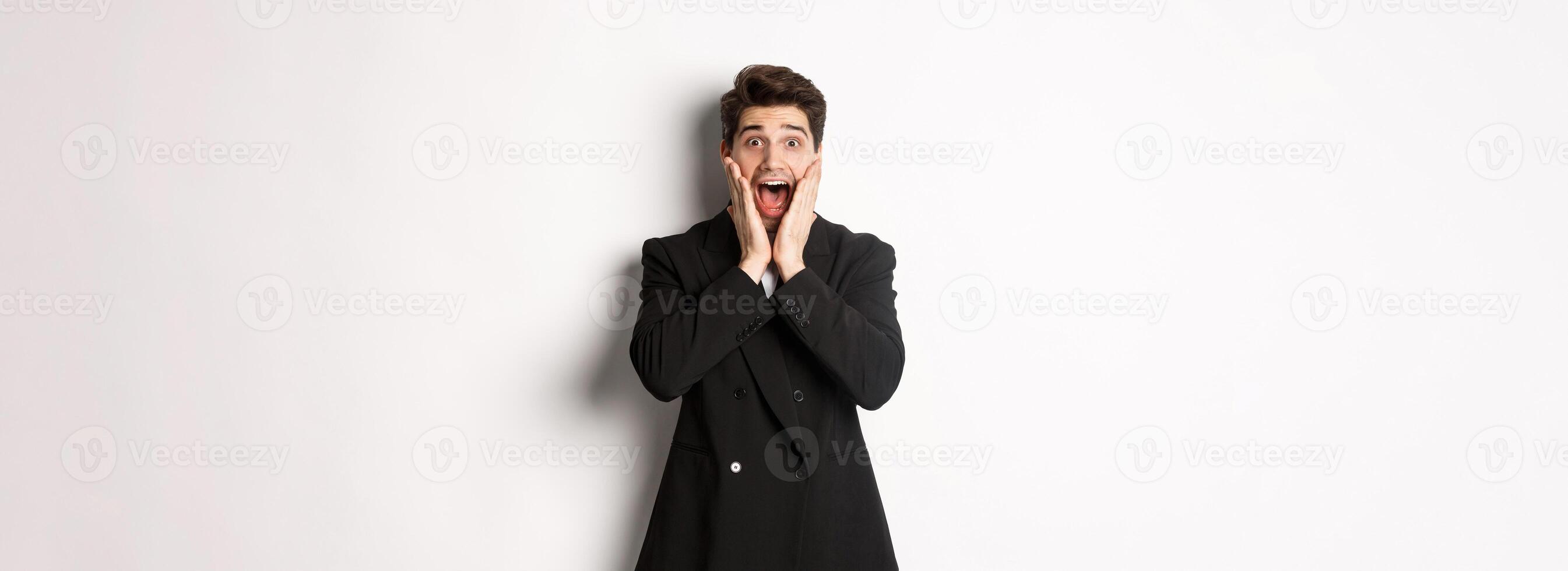 Portrait of surprised handsome businessman in suit, reacting to holidays promo, looking amazed at camera, standing over white background photo
