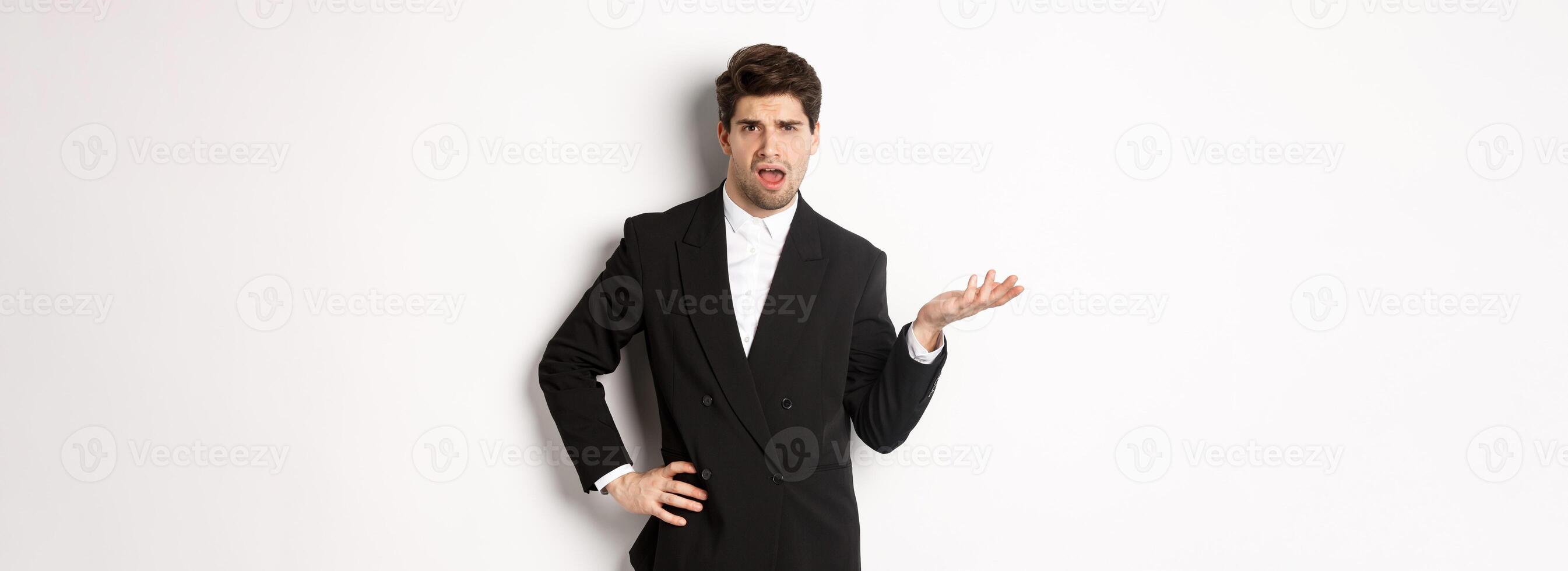 Portrait of arrogant man in black suit, looking confused and disappointed, complaining about something strange, standing over white background photo