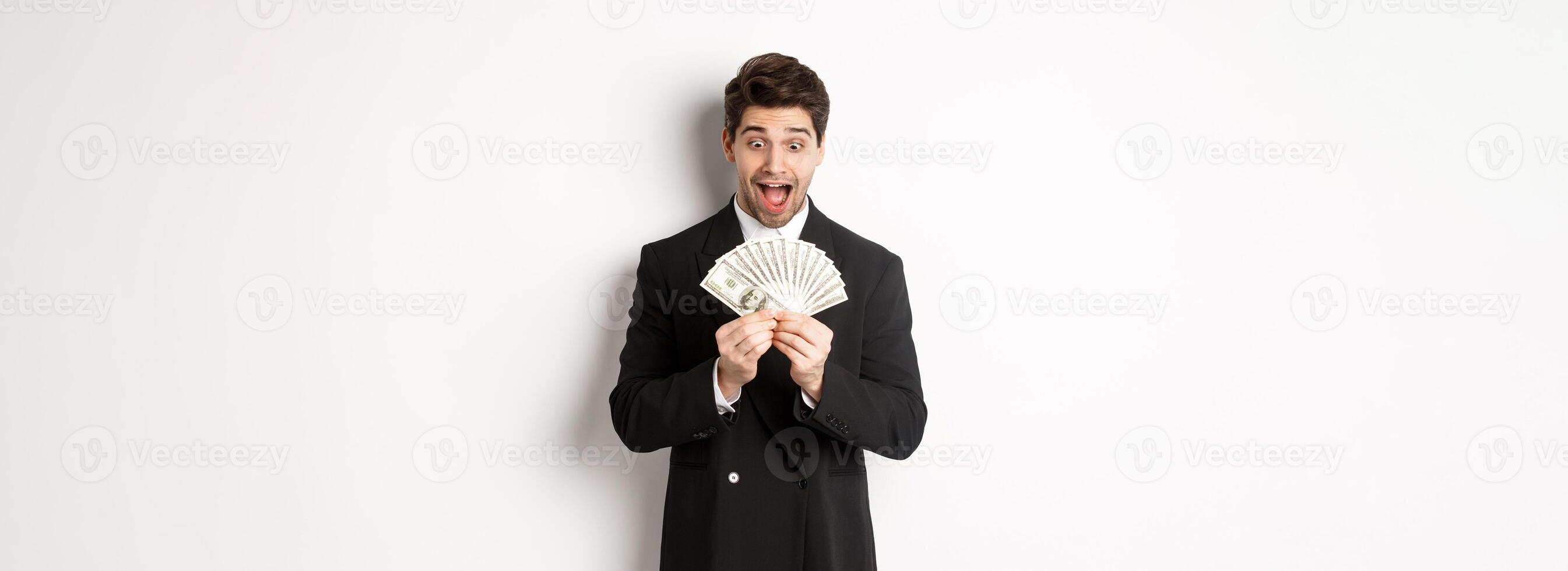 Image of handsome bearded guy in black suit, looking at money with excitement, standing over white background photo