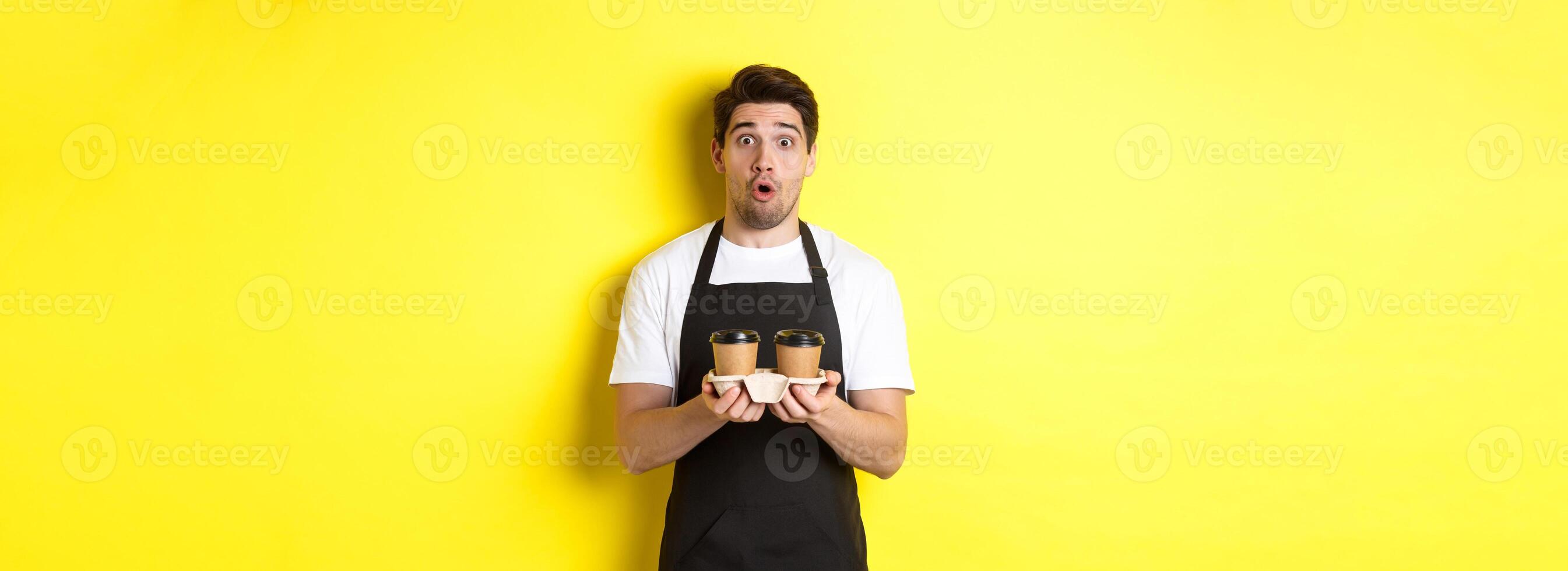 Barista serving coffee, looking surprised at camera, wearing black apron, standing against yellow background photo