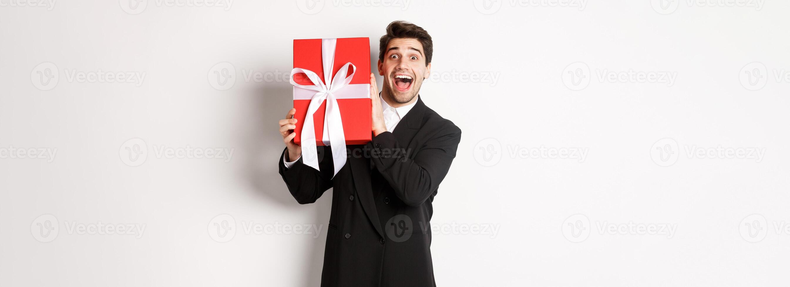 Image of happy good-looking man in black suit, holding box with christmas gift and smiling, standing against white background photo